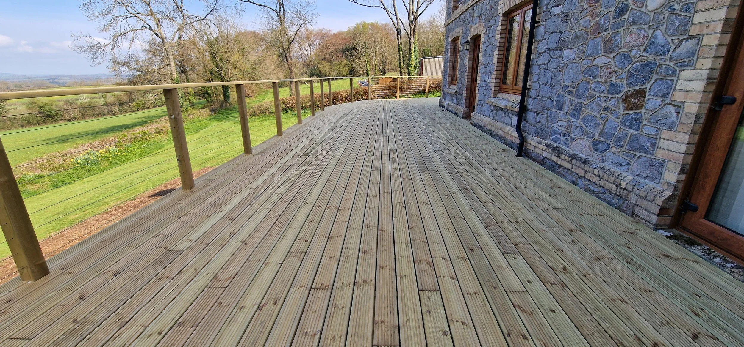 New wooden deck attached to brick house, overlooking a grassy landscape with trees and cloudy sky.