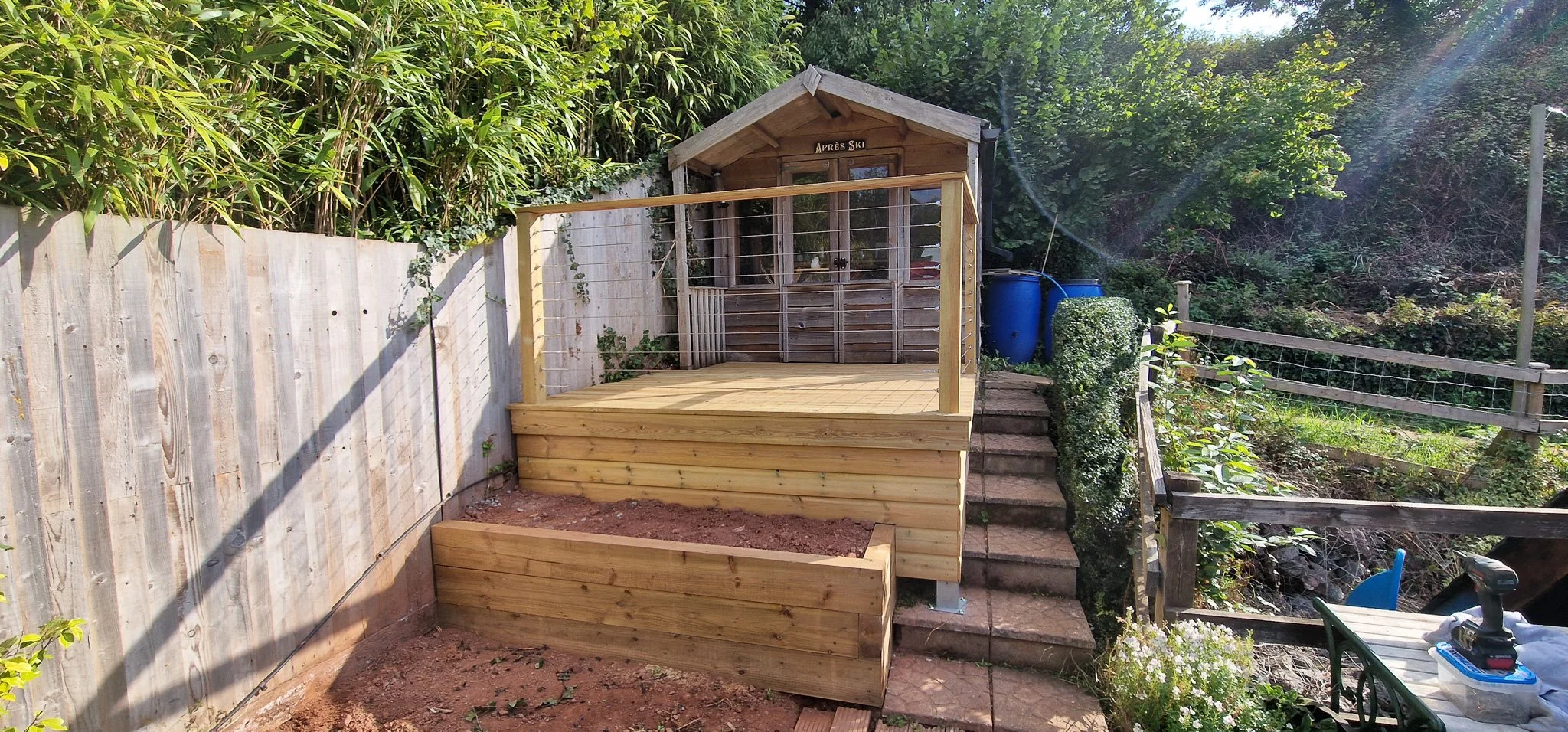 New wooden deck with a small storage shed behind it, stairs leading up to the shed, and blue rain barrels nearby, surrounded by lush greenery.
