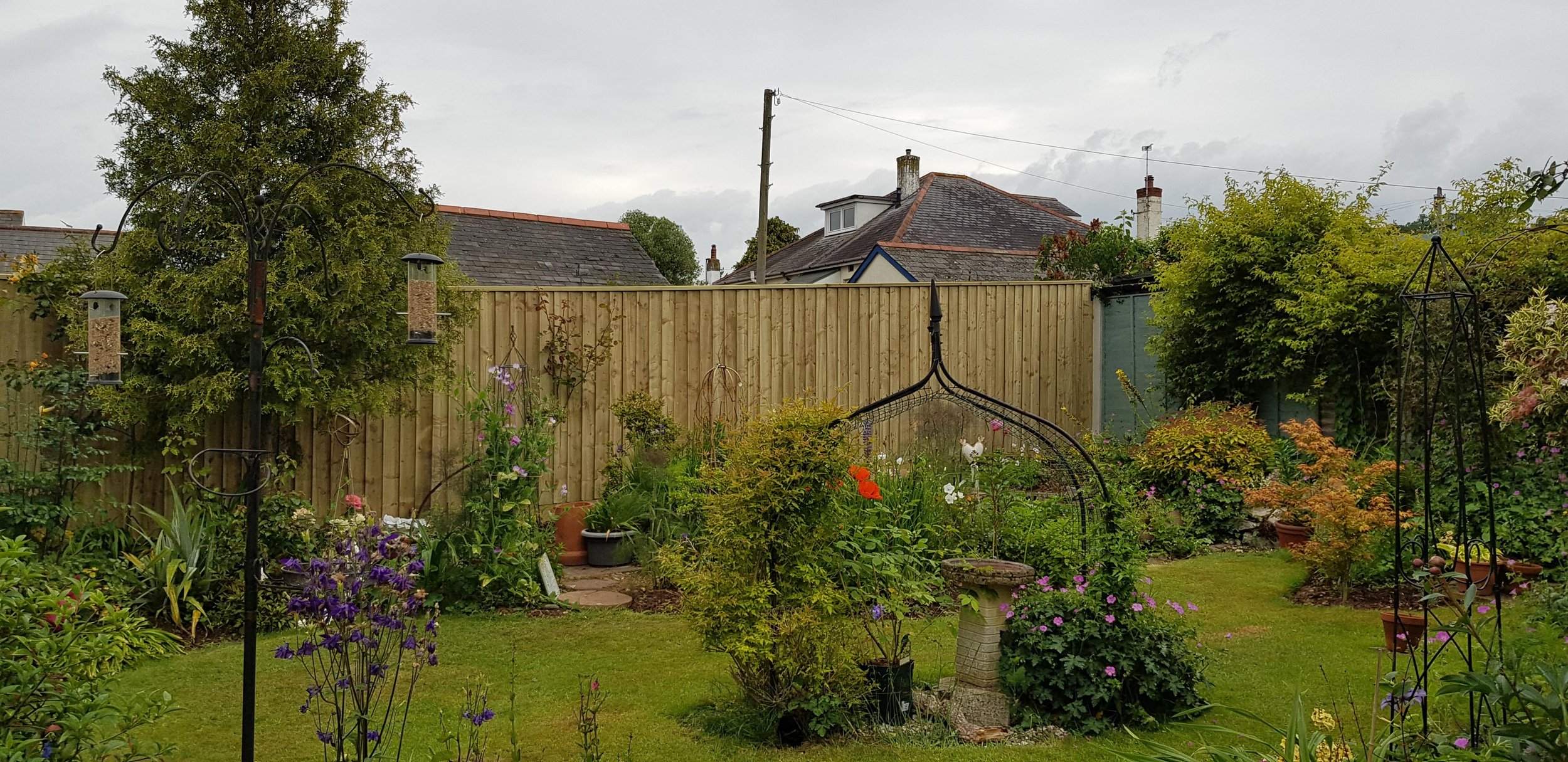 A neatly maintained garden with colorful flowers, potted plants, garden ornaments, bird feeders, and a wooden fence in the background, with neighboring rooftops and cloudy sky overhead.