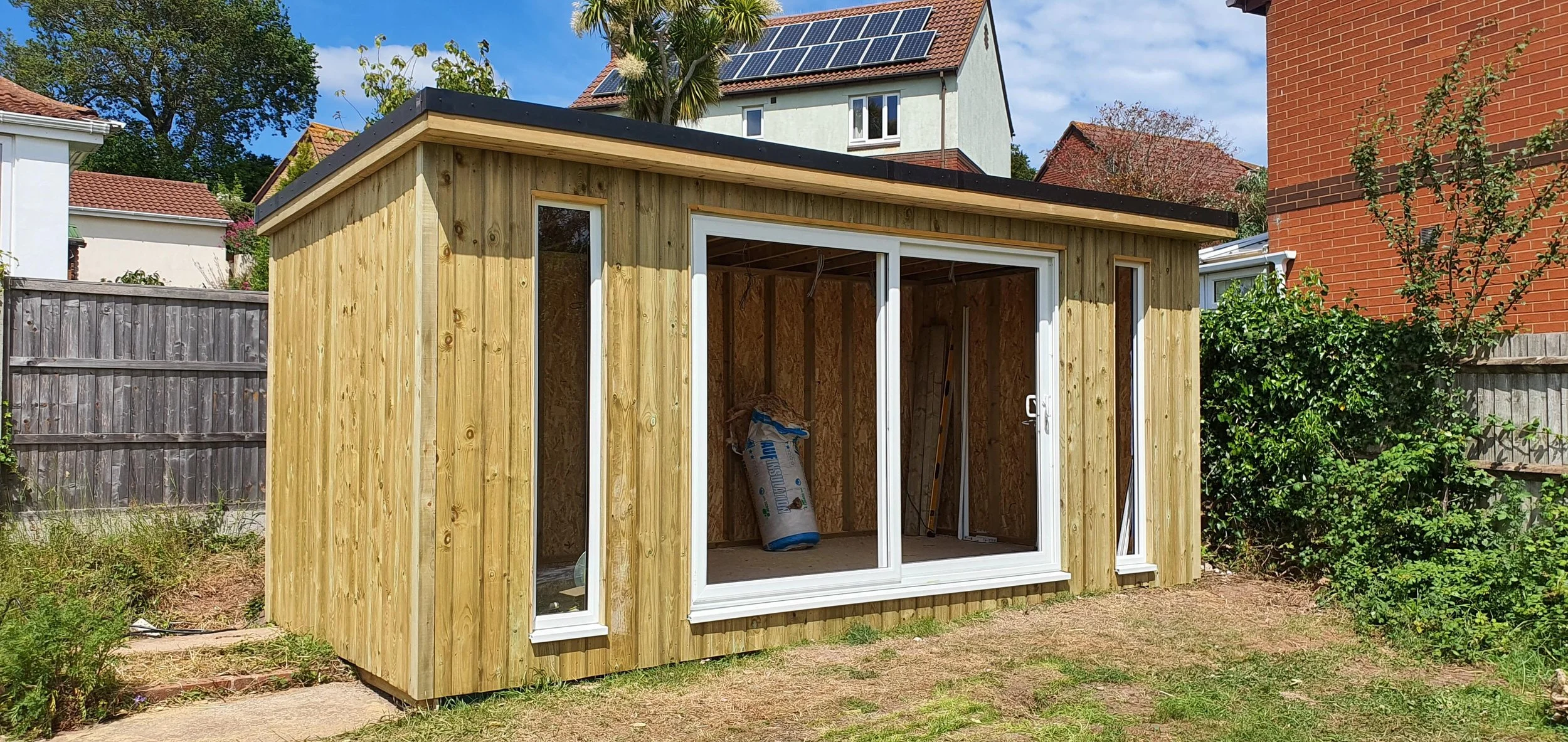 Wooden garden shed with large sliding glass doors, situated in a backyard with neighboring houses and fenced yard.