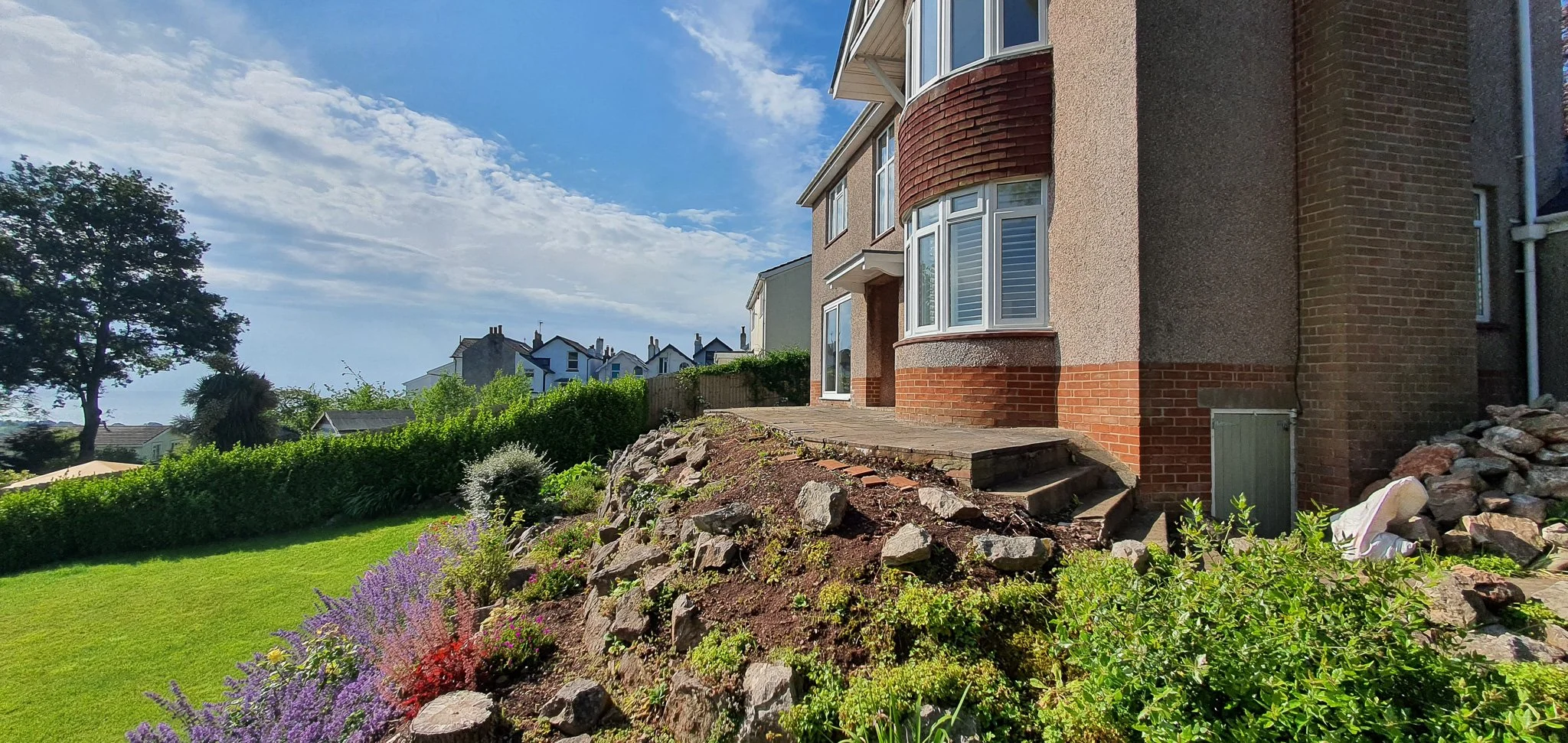 Large brick and stucco house with bay windows on a sunny day, surrounded by a landscaped garden with colorful flowers, rocks, and green grass.