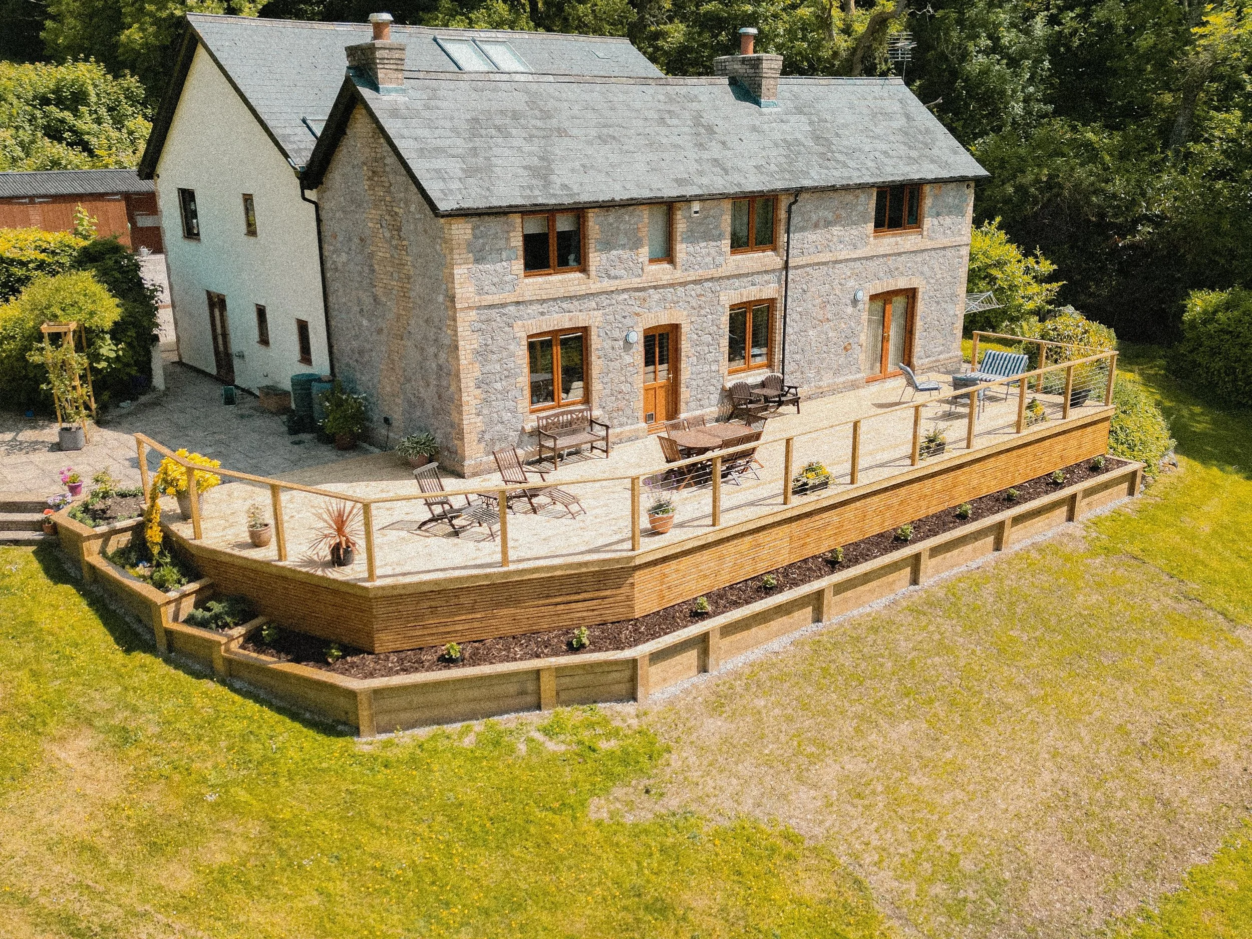A large stone and brick house with a spacious wooden deck balcony in the backyard. The deck has outdoor furniture including chairs, a table, and potted plants, surrounded by a wooden railing. The backyard is grassy with garden beds and greenery surro