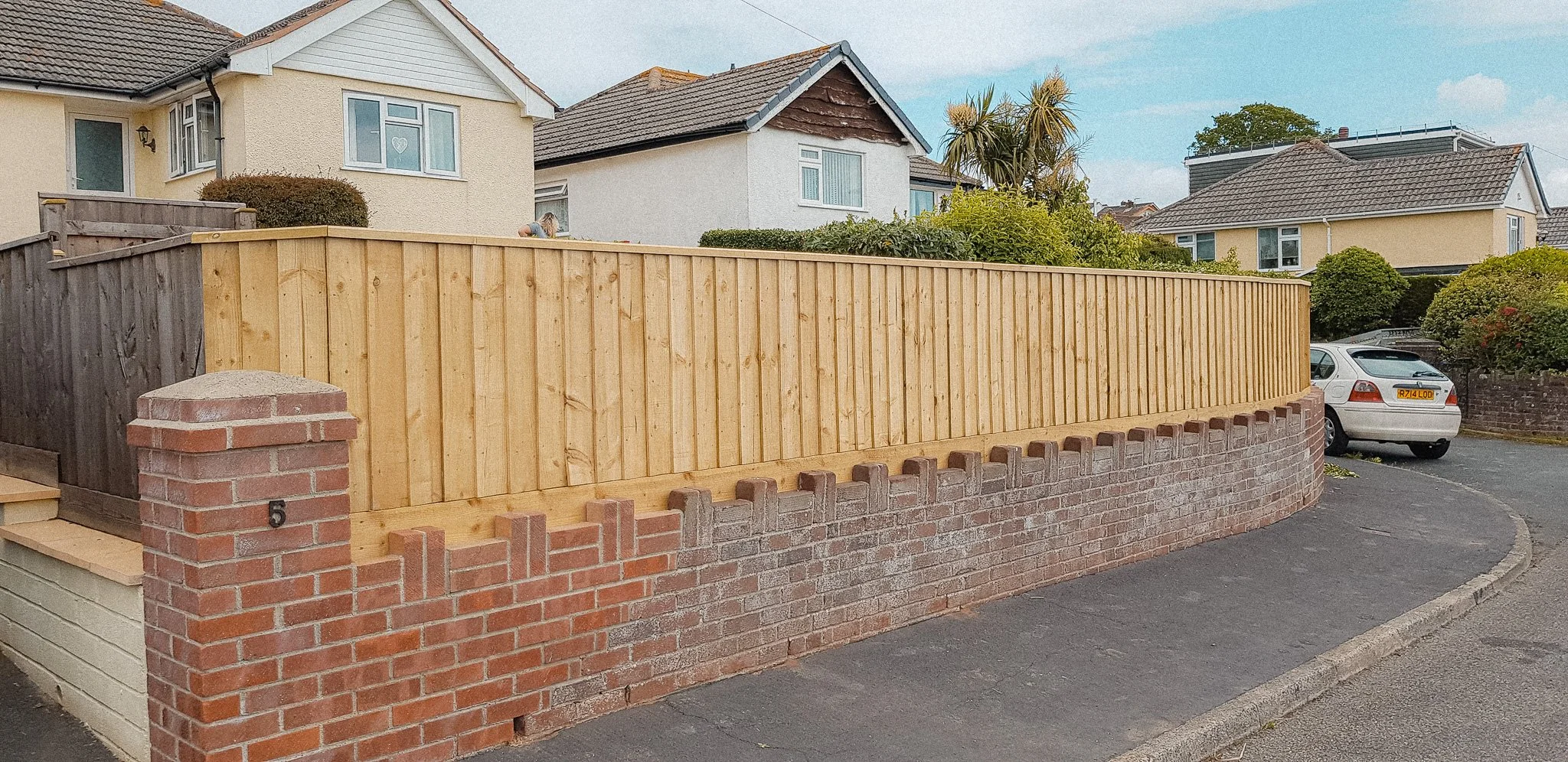 A new wooden fence installed along a brick wall boundary in front of residential houses on a street, with a parked silver car nearby.