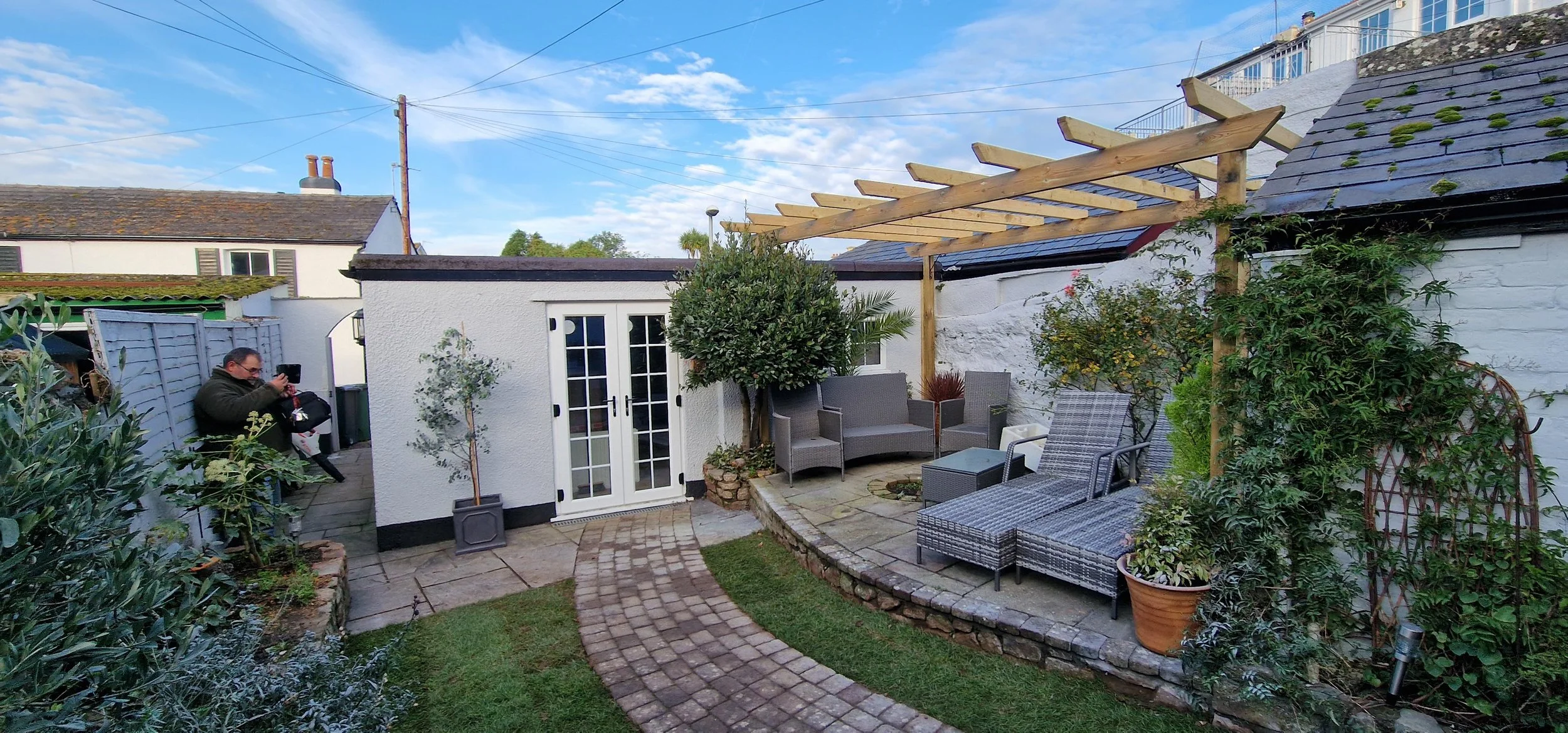 Backyard patio with a curved brick pathway, outdoor furniture including a wicker lounge chair, a small sofa, and a glass-top table, potted plants, and a wooden pergola frame without a cover. A small white building with glass double doors is also visi