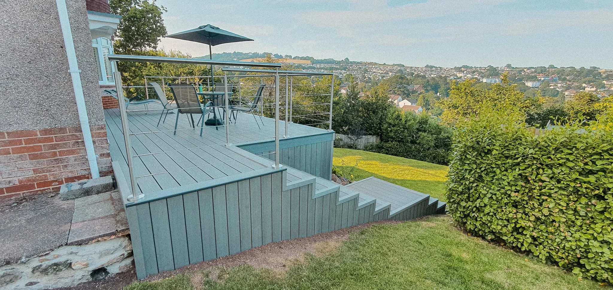 View of a backyard deck with outdoor furniture, staircase, and railing, overlooking a green lawn, trees, and a distant neighborhood on a clear day.