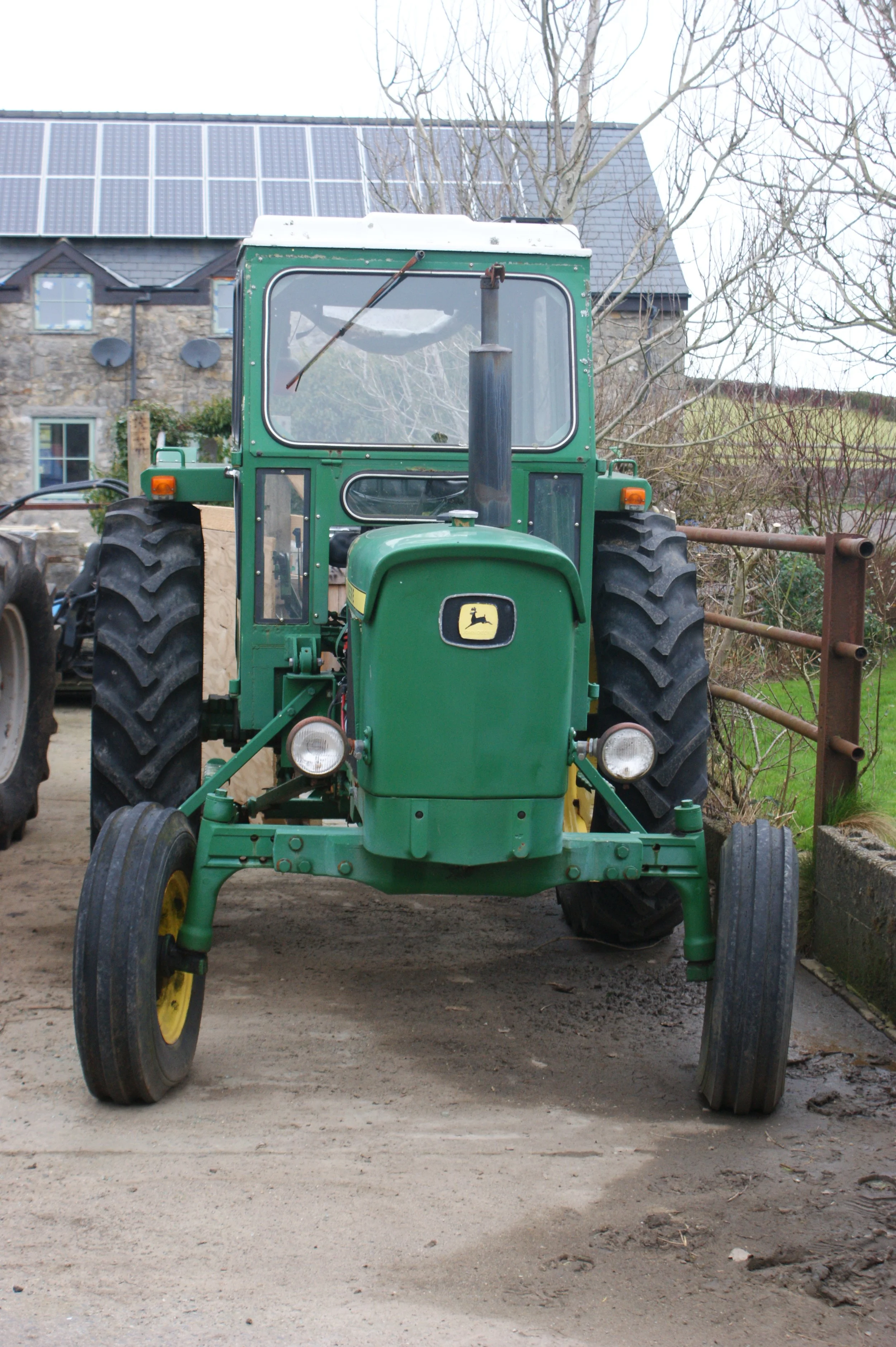 Front view of a green John Deere tractor on a dirt surface, with a stone house and solar panels on the roof in the background, and leafless trees nearby.