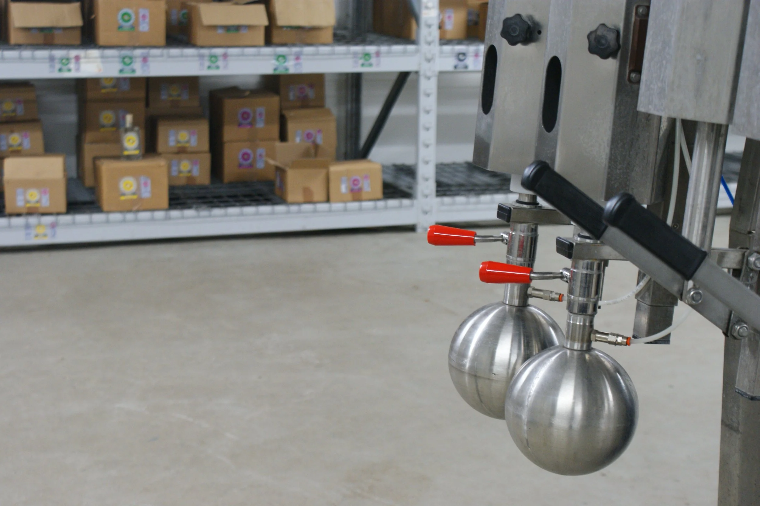 Industrial machinery with two stainless steel spherical components and red-handled valves, in a warehouse with shelves of cardboard boxes.