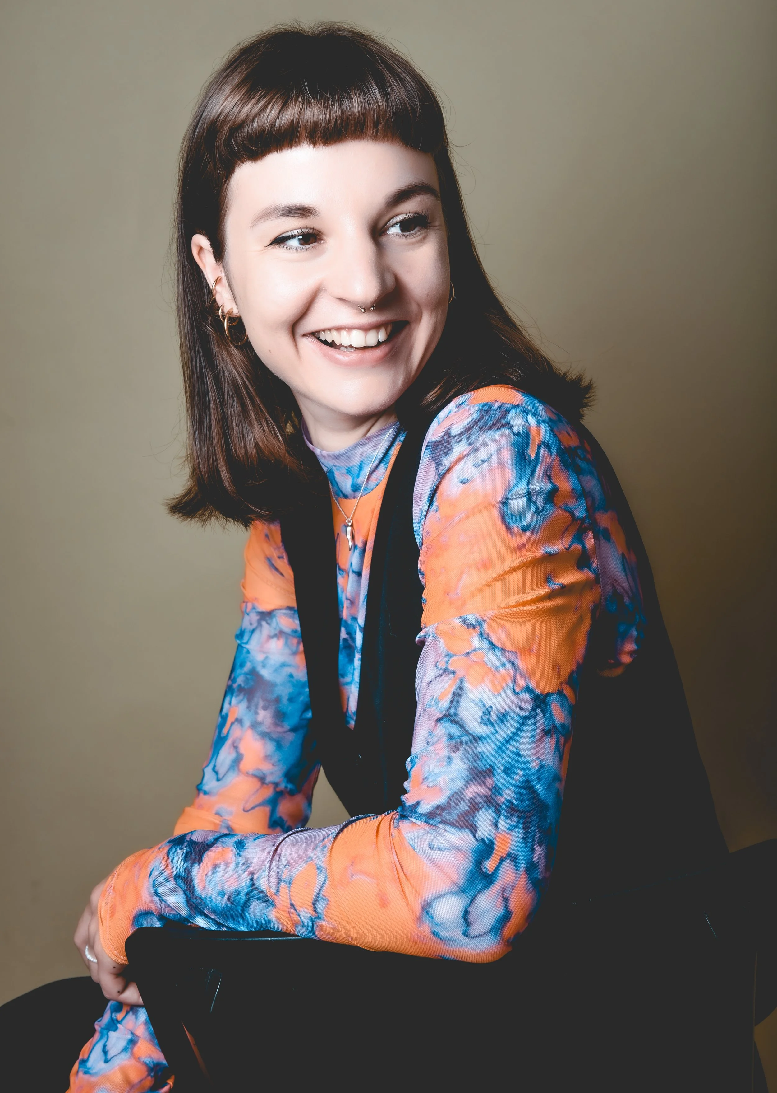 Junge Frau mit braunen Haaren, Kurzhaarschnitt, lächelnd, trägt ein buntes, langärmeliges Shirt mit Orange, Blau und Lila Mustern, vor einem beigefarbenen Hintergrund.
