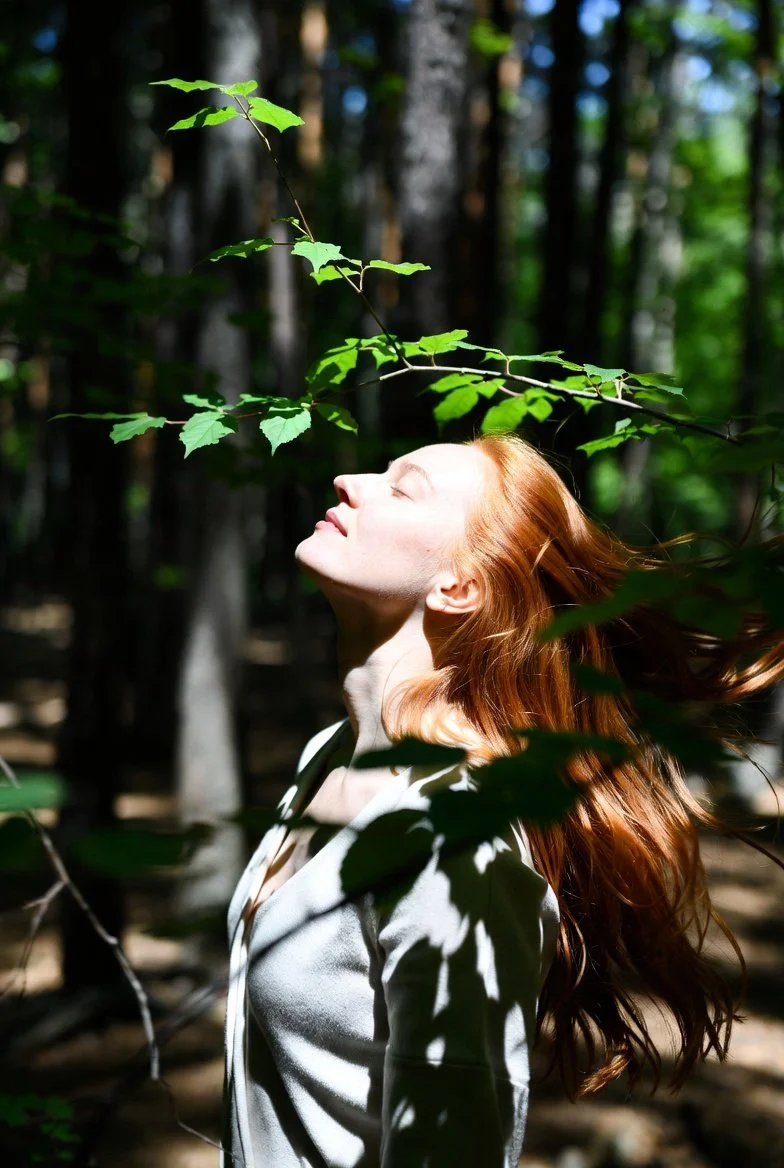 A woman with long red hair and closed eyes stands in a wooded forest, basking in sunlight filtering through green leaves.