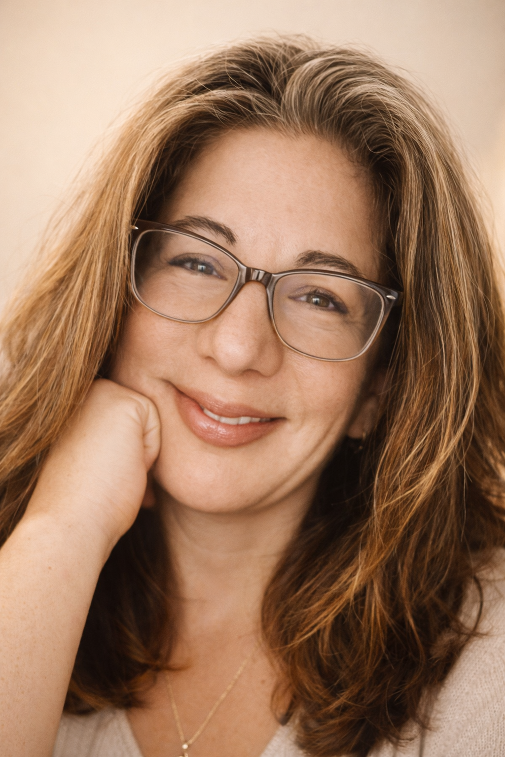 Close-up of the creator- a woman with long, wavy auburn hair wearing glasses, smiling gently, resting her face on her hand.
