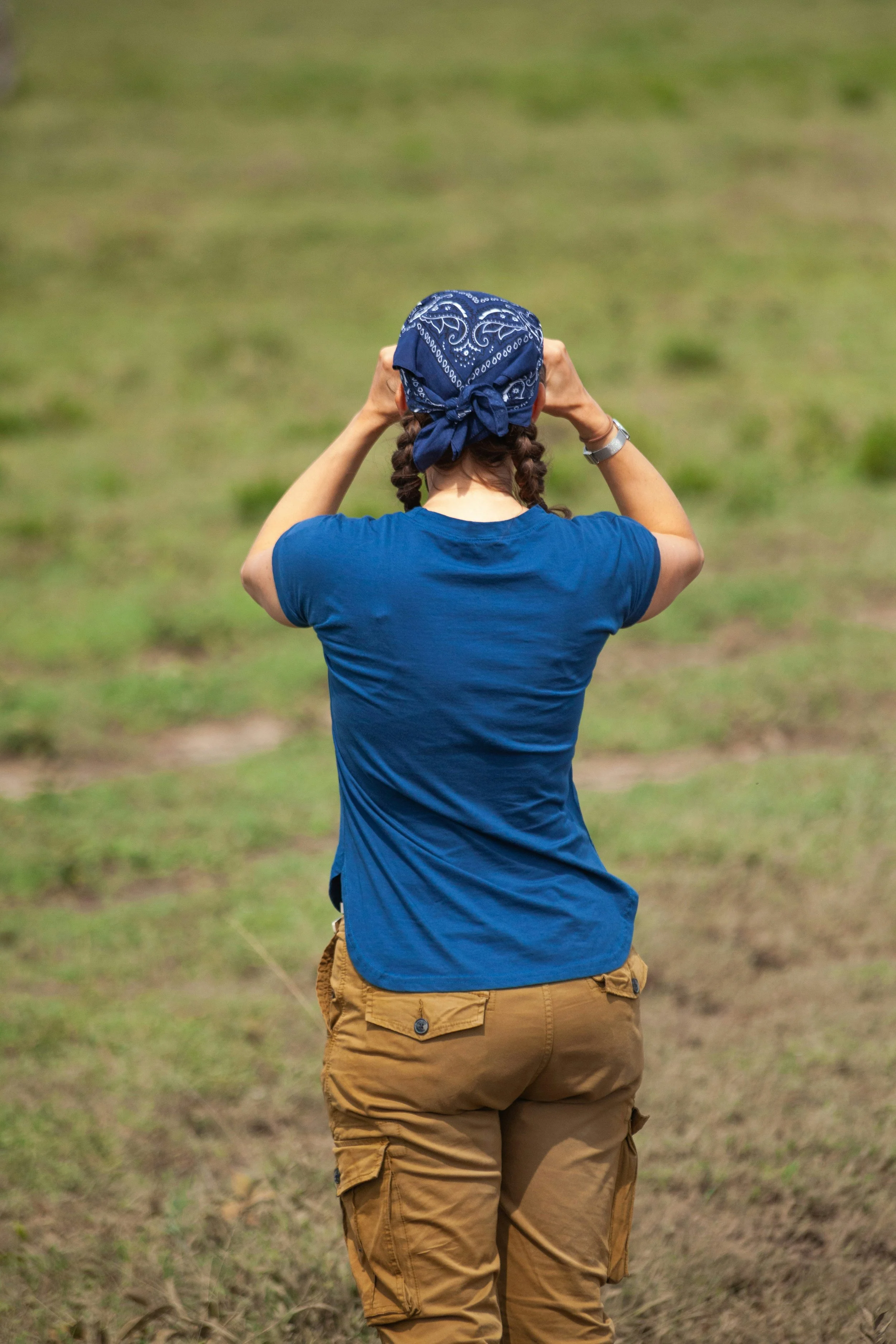 Person with braided hair wearing a blue bandana, blue t-shirt, and tan cargo shorts, standing in an open grassy field.