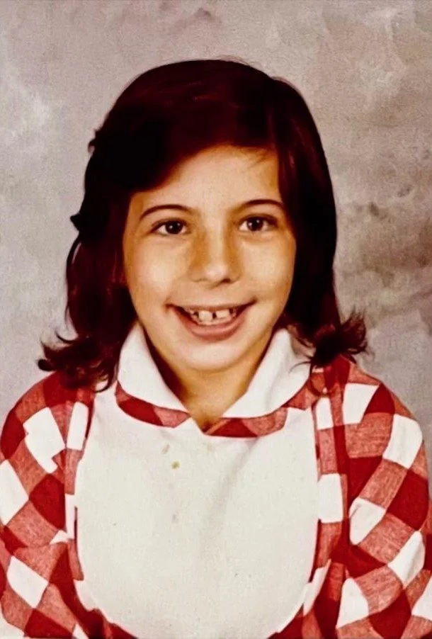 A young girl with dark hair, buck teeth, and a big smile, wearing a red and white checkered shirt.