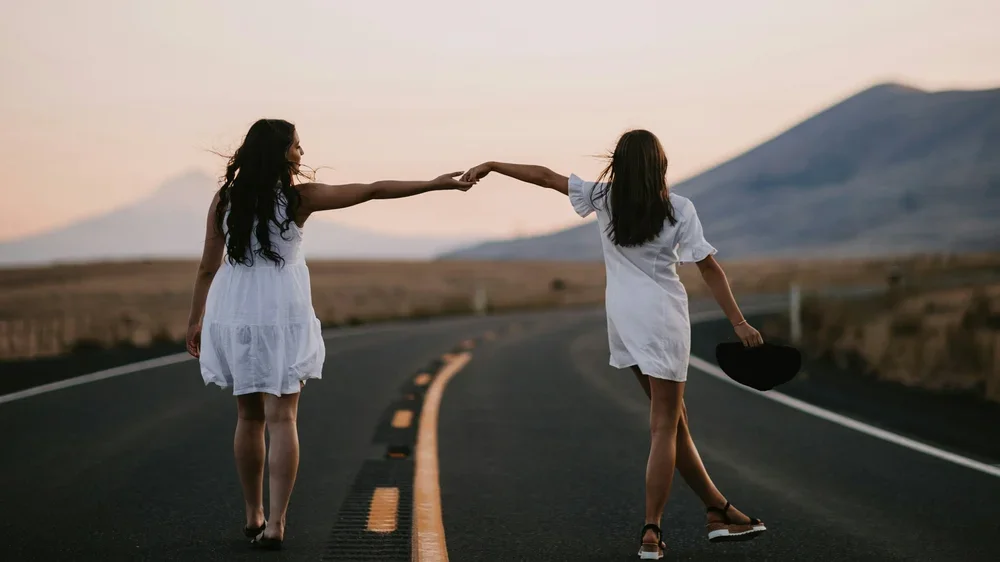 Two women in white dresses holding hands while walking away on an empty road during sunset, with mountains in the background.