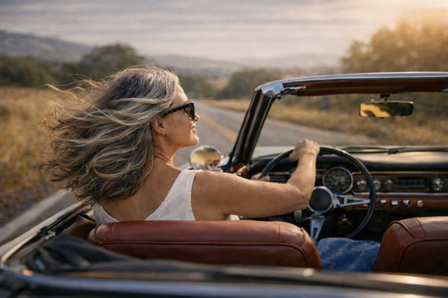 A woman with long gray hair driving a convertible car on an open road during sunset.
