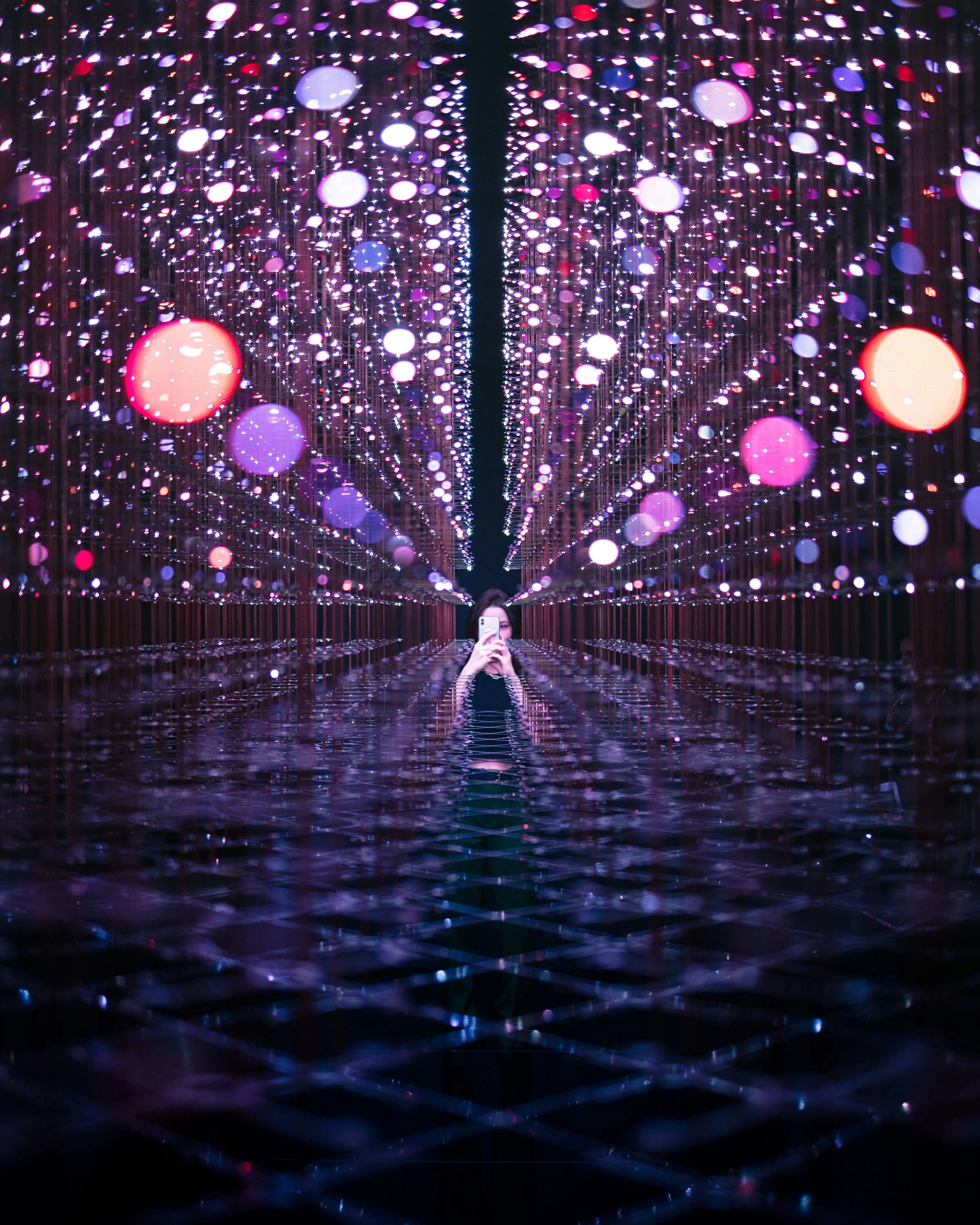A person is taking a photo of a ceiling with colorful, circular LED lights in an infinity mirror exhibit.