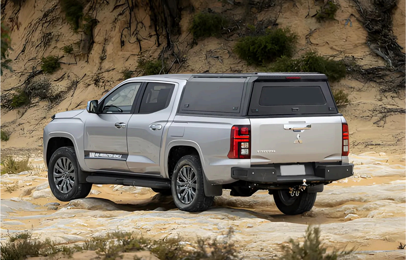 Silver pickup truck with black camper shell driving on rocky, sandy terrain in desert landscape.