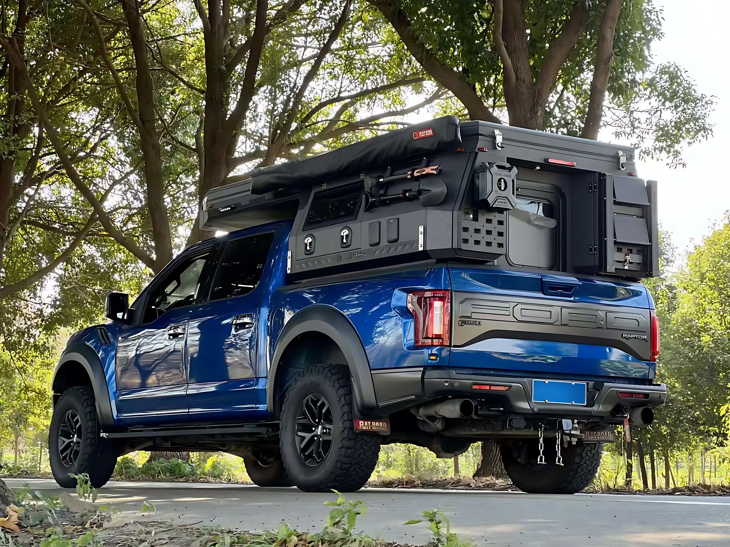 A blue Ford F-150 pickup truck with a large, black roof-mounted camper and outdoor camping gear in a wooded area.