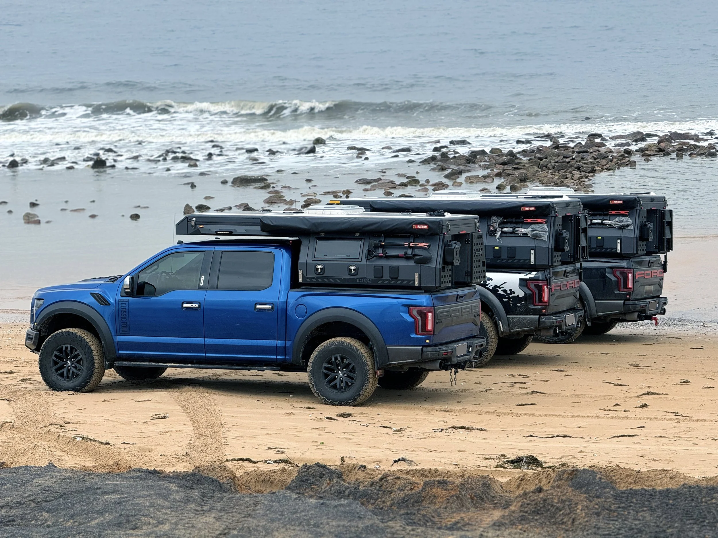 Three blue pickup trucks with gear mounted in the truck beds are parked on a beach with sandy and rocky areas, facing the ocean.