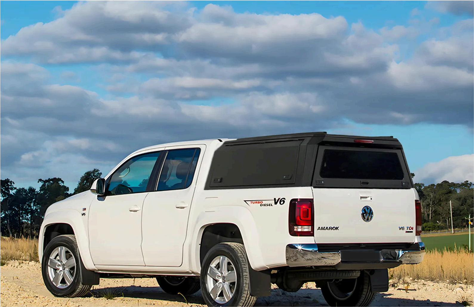 White Volkswagen Amarok pickup truck with a black canopy on a dirt road during daytime, under a partly cloudy sky.