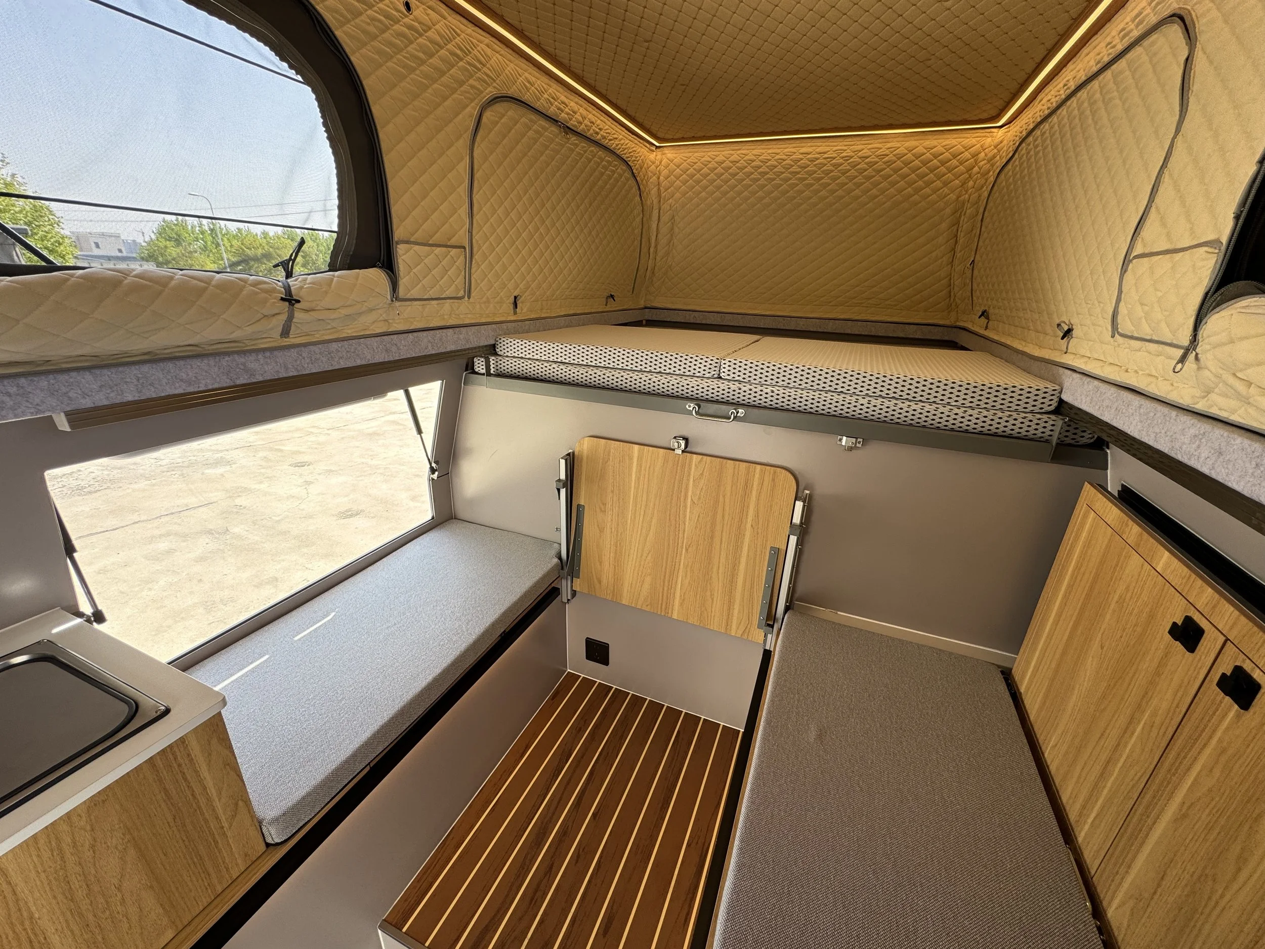 Interior of a compact camper van with wooden cabinetry, padded sleeping area with yellow quilted walls, and large side windows.