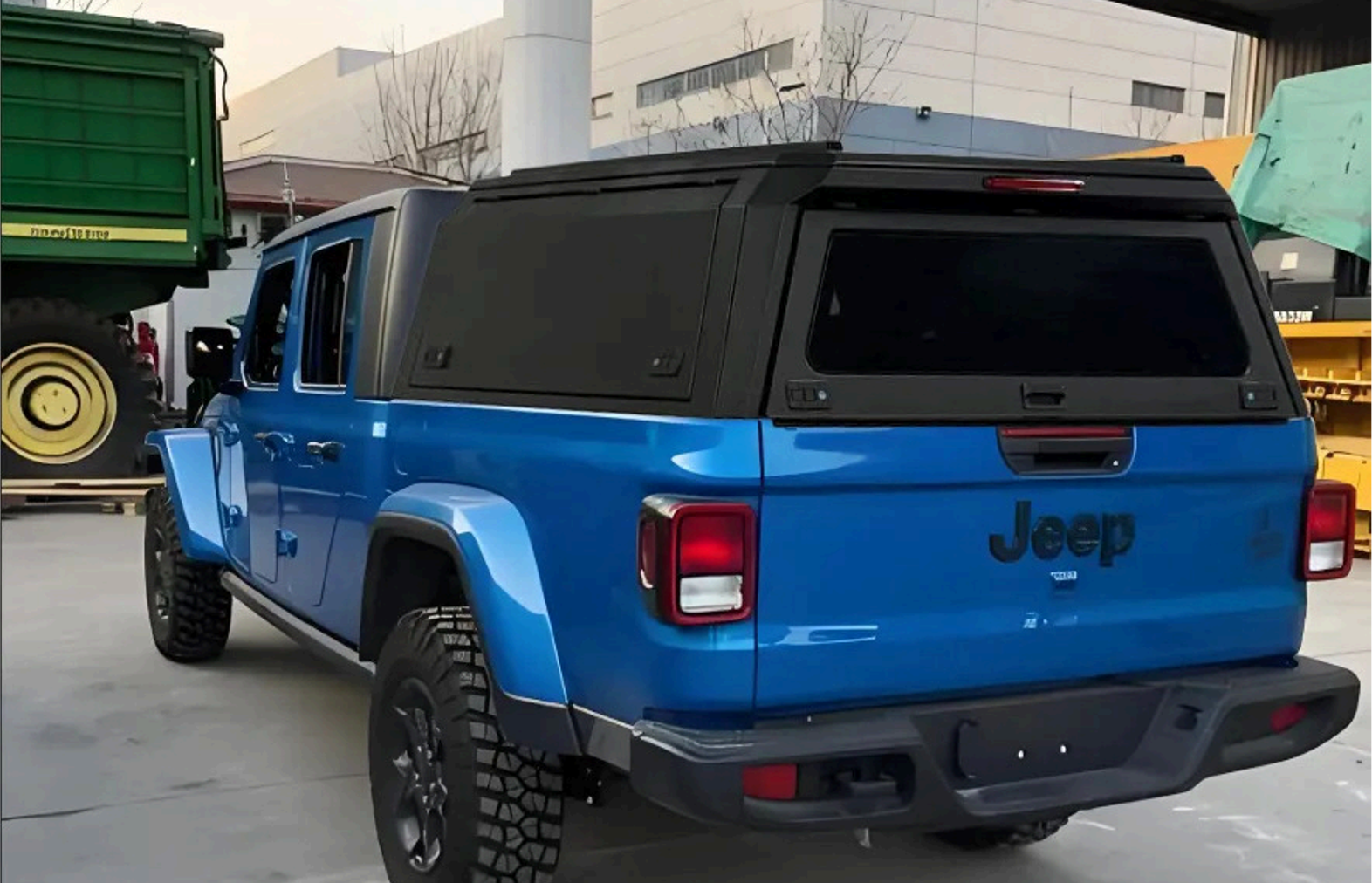 Blue Jeep with black rooftop storage in a parking lot, surrounded by industrial equipment and buildings.