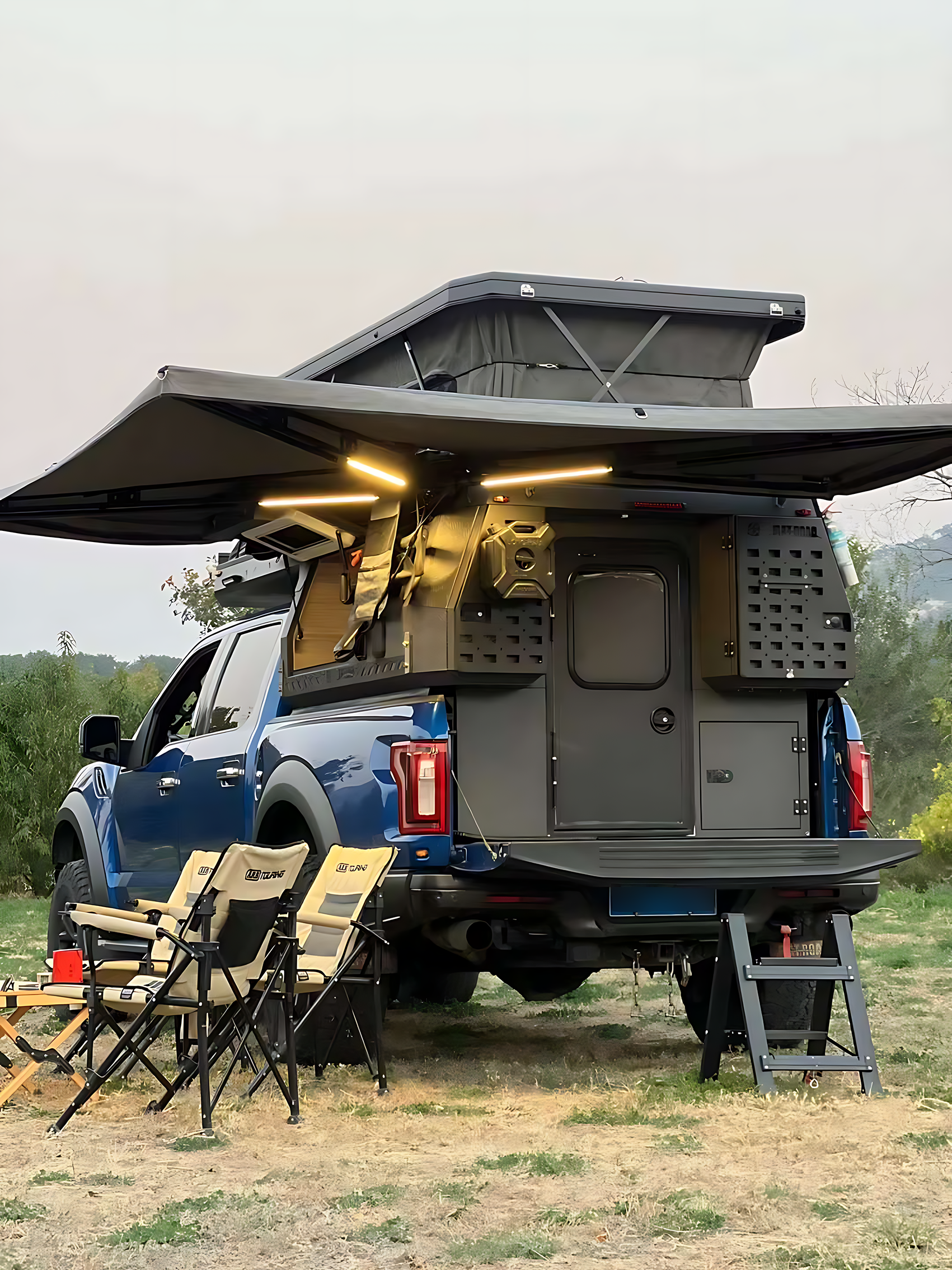 Blue truck with a camper setup, open side awning, outdoor chairs, and stairs leading to the camper door in a grassy outdoor setting.