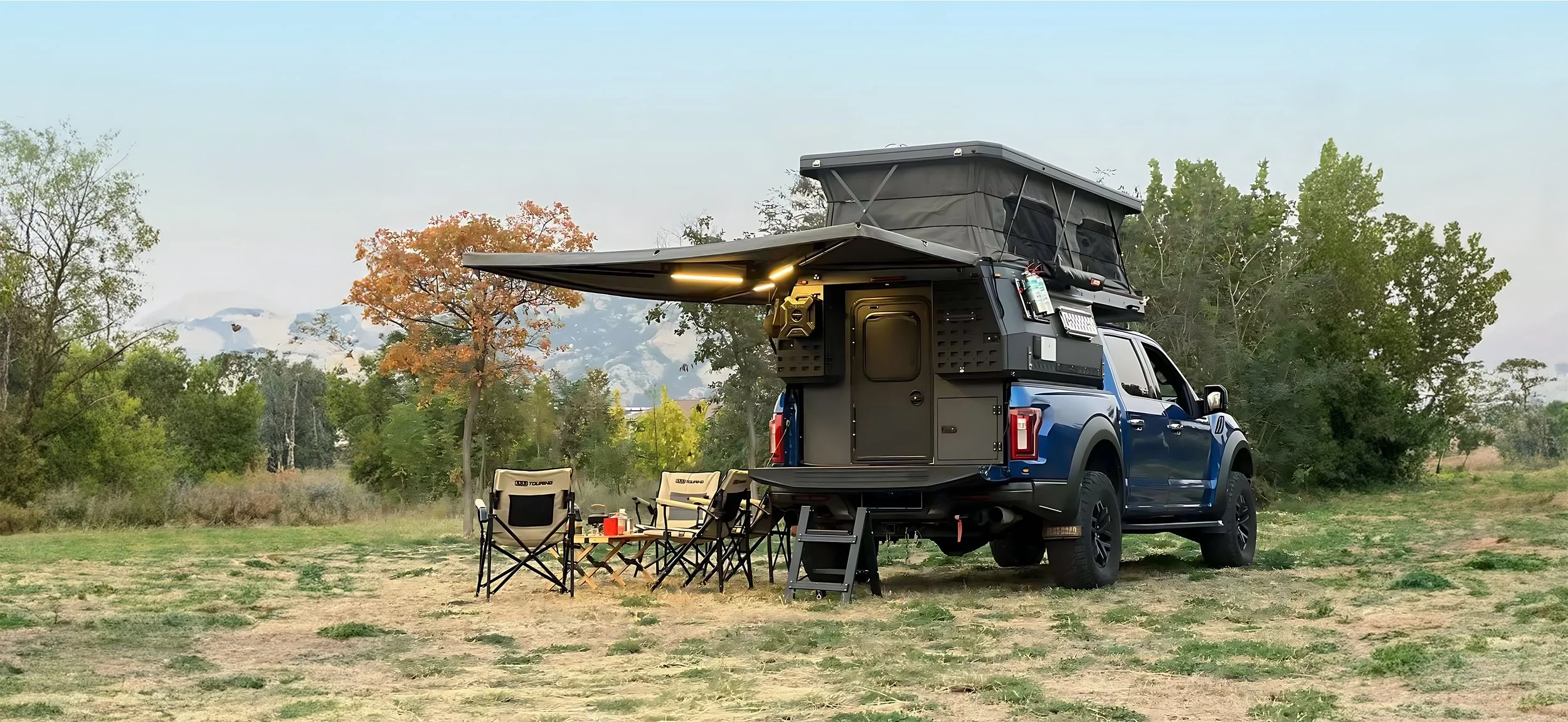 Blue pickup truck with a rooftop tent set up in a grassy outdoor area, surrounded by trees and mountains in the background, with camping chairs and a small table nearby.