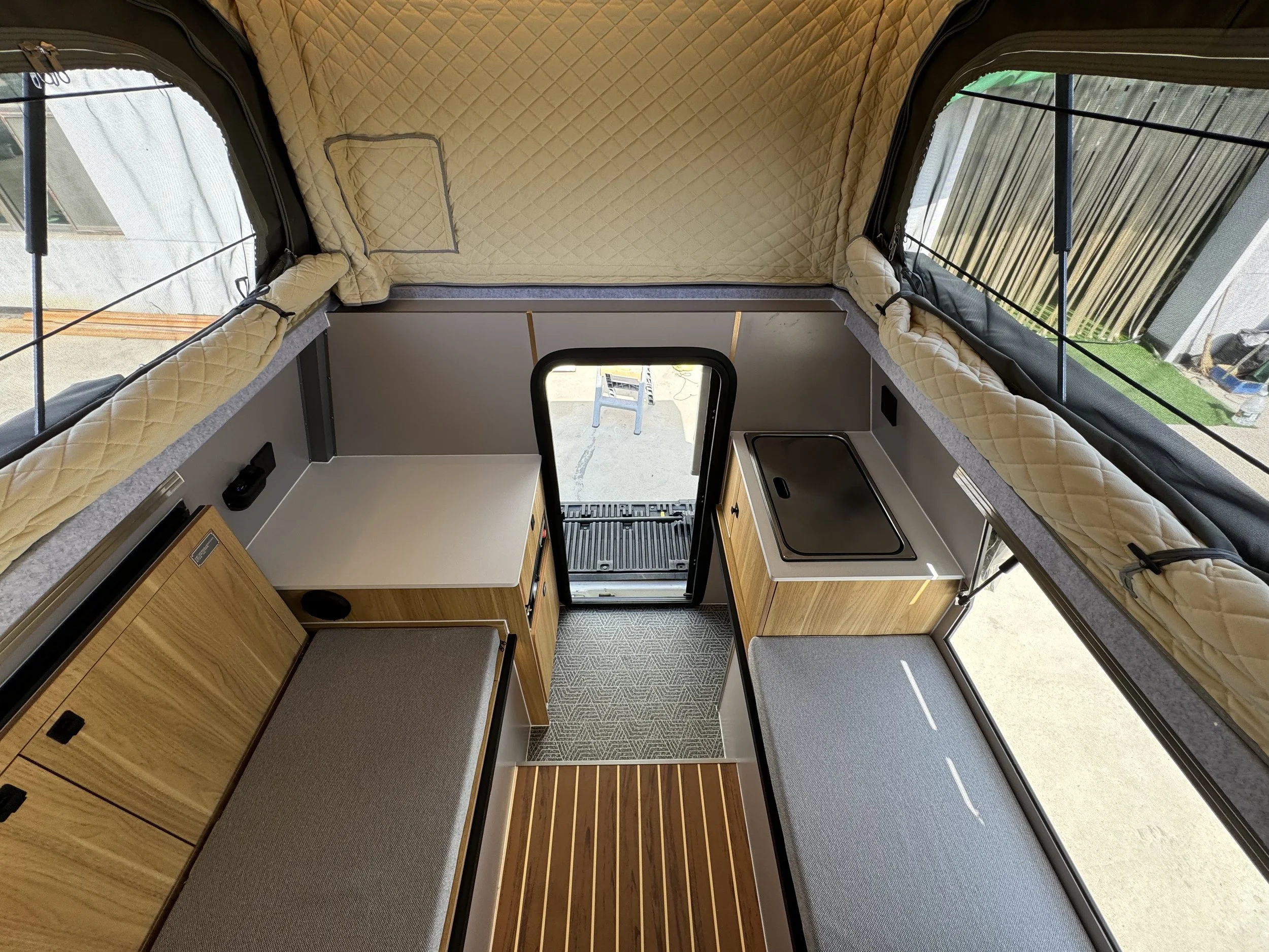 Interior of a camper with wooden cabinets, grey cushions, a small kitchenette with a cover, and large windows with quilted beige curtains.