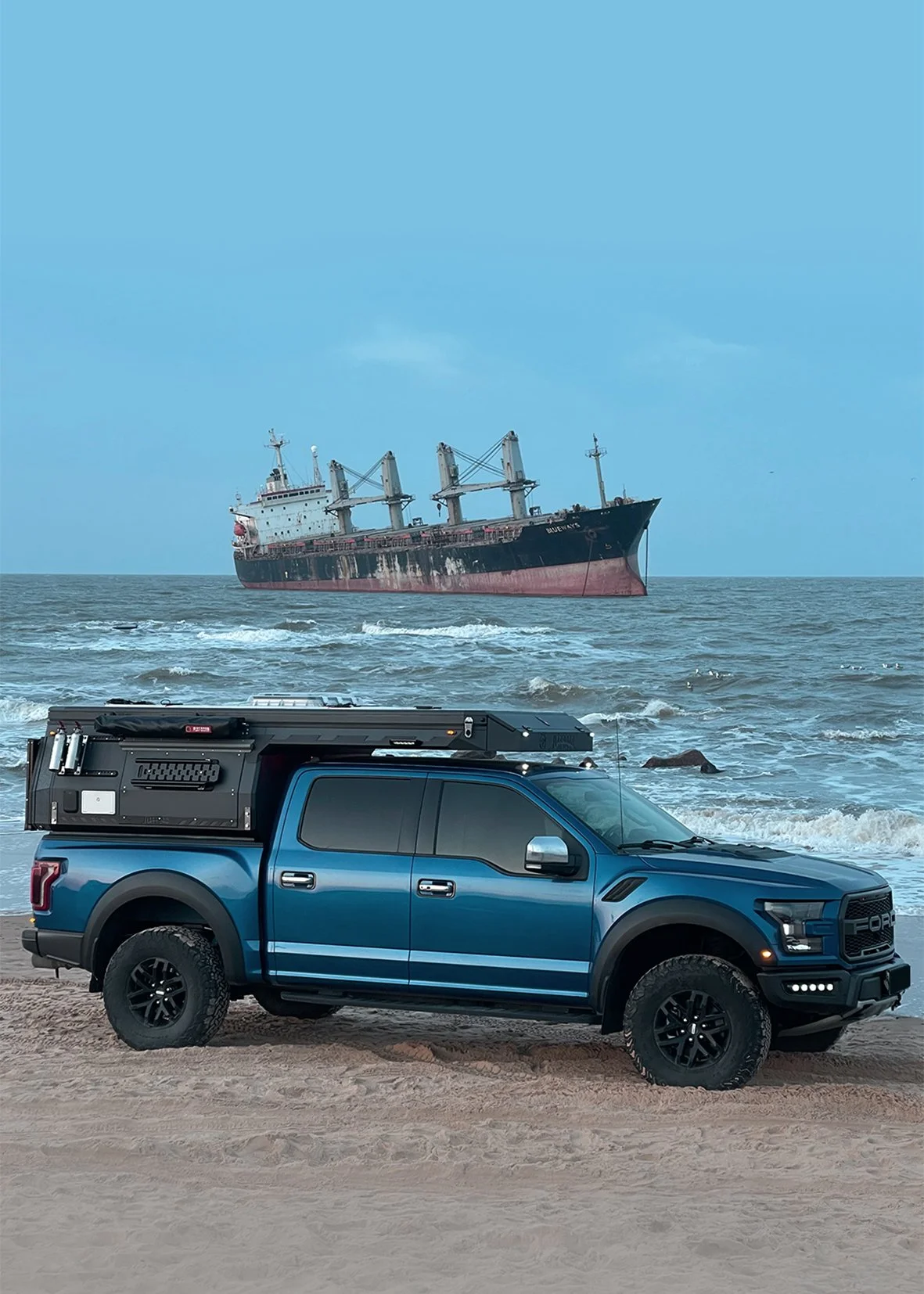 A large cargo ship at sea with a black and red hull, viewed from a beach where a blue pickup truck with equipment on top is parked.