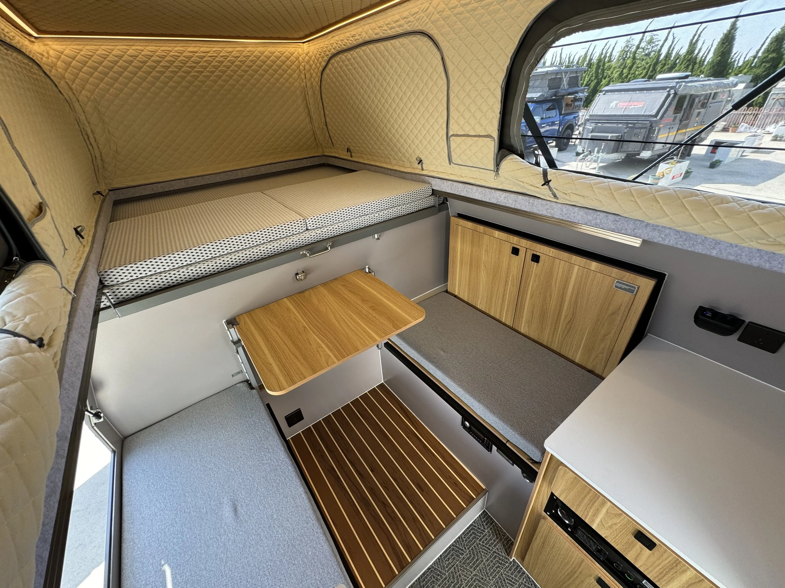 Interior of a camper van with a sitting area, a bed above, and a window showing parked RVs outside.
