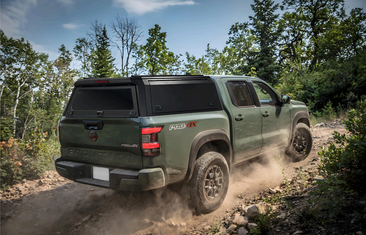 Green Nissan Frontier PRO-4X pickup truck driving off-road on a dirt trail surrounded by green trees and bushes.