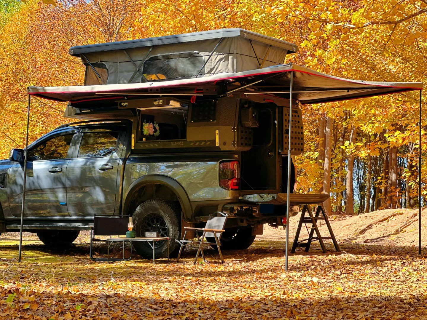 A camping trailer attached to a pickup truck with an outdoor setup including folding chairs, a small table, and camping gear, surrounded by autumn trees with yellow and orange leaves.