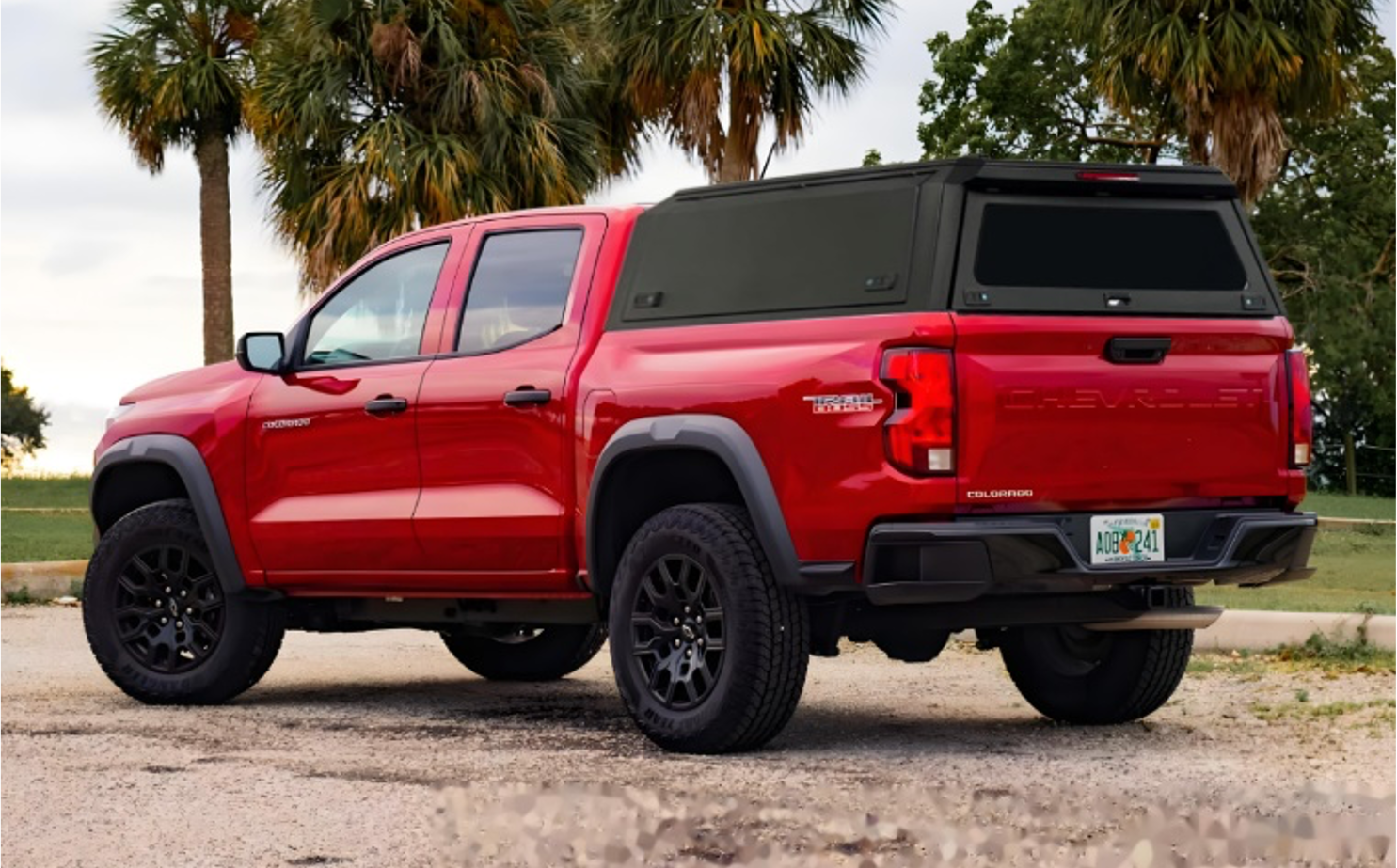 Red Chevrolet Colorado pickup truck parked on a gravel surface with palm trees and green trees in the background.