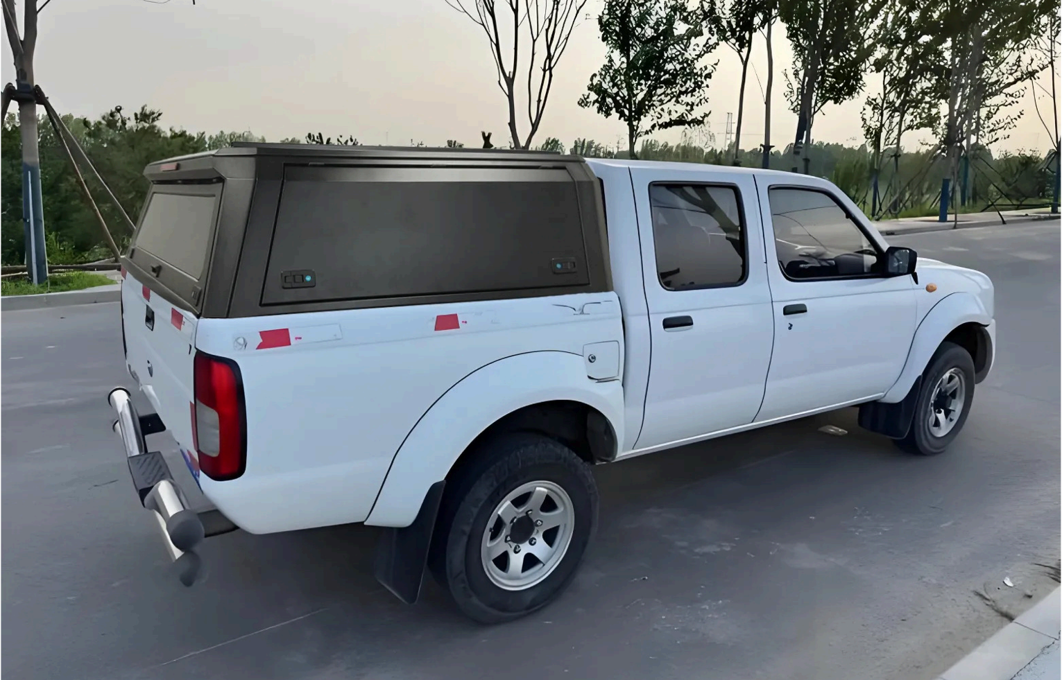 White pickup truck with black canopy parked on a paved area, trees and overcast sky in the background.