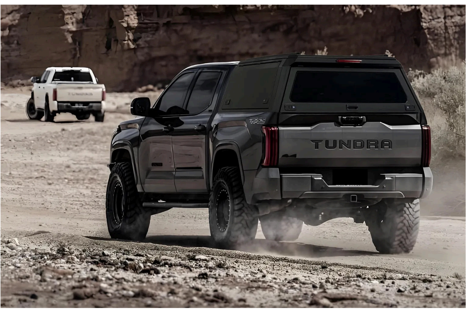 A black Toyota Tundra pickup truck driving on a dusty, rocky terrain with dust being kicked up by its tires, in a desert landscape with rock formations in the background.