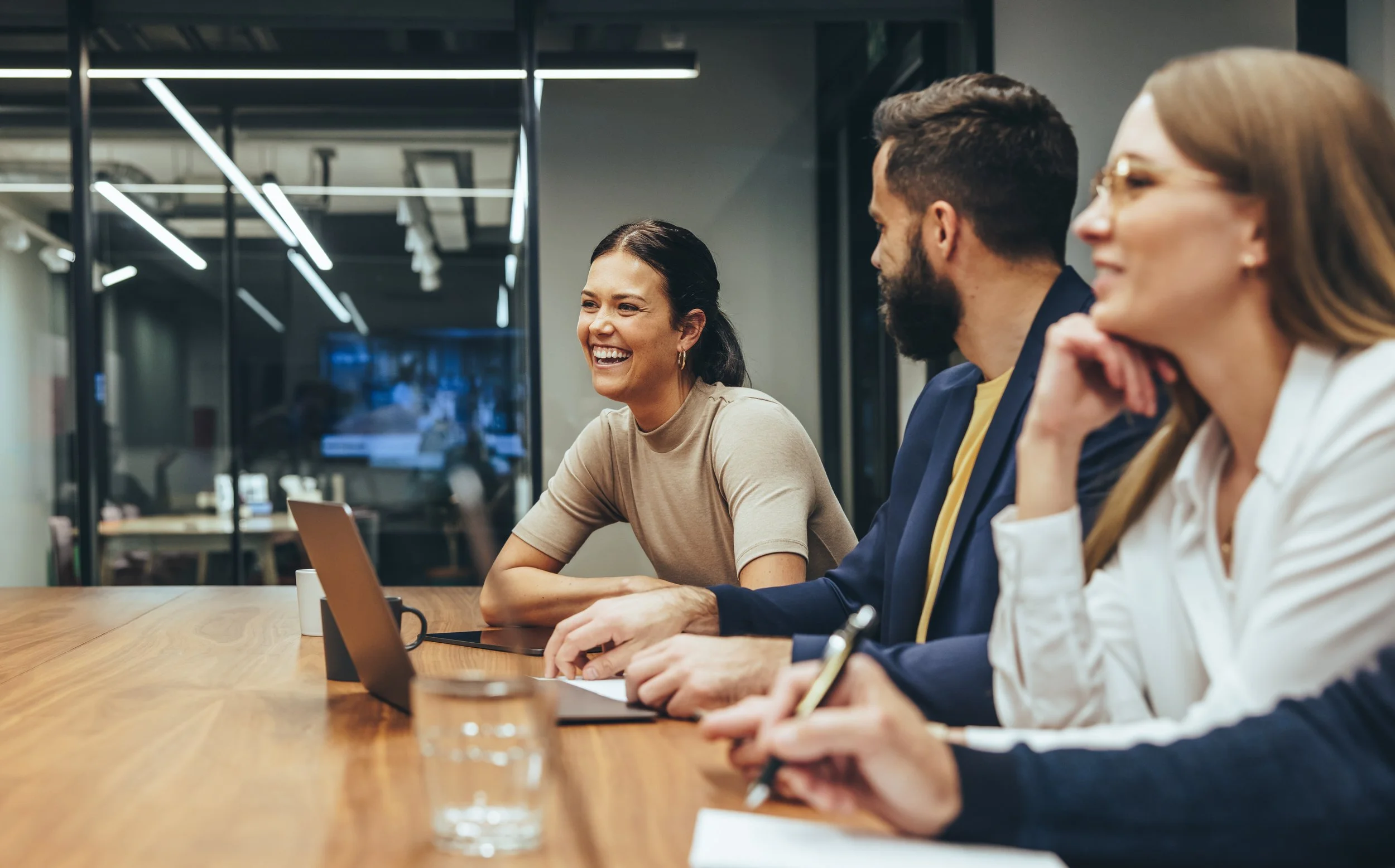 3 office staff in a meeting room, they sit on the right hand side of the table, female with glasses, male with beard and woman with pony tail. All are smiling. 