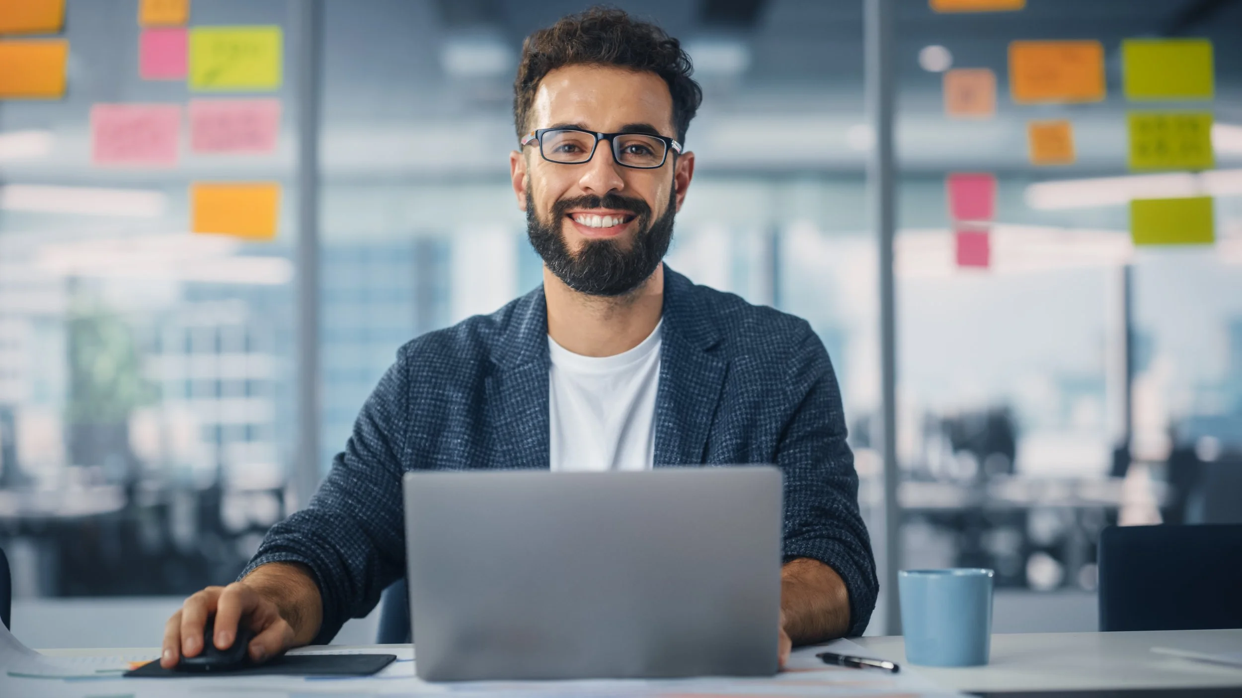 A smiling man with glasses, a beard, and dark curly hair working at a desk in an office with colorful sticky notes on the glass wall behind him, using a laptop and a mouse.