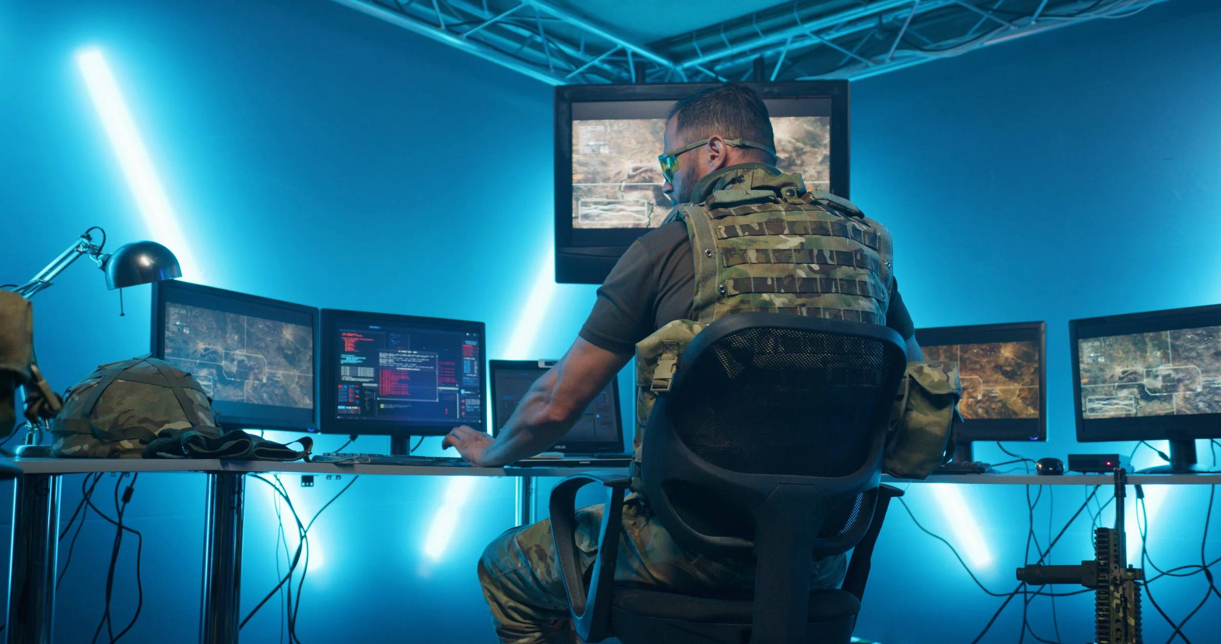 A man in military camouflage attire sitting at a desk with multiple computer screens, monitoring a video game or simulation, in a high-tech room with blue lighting.