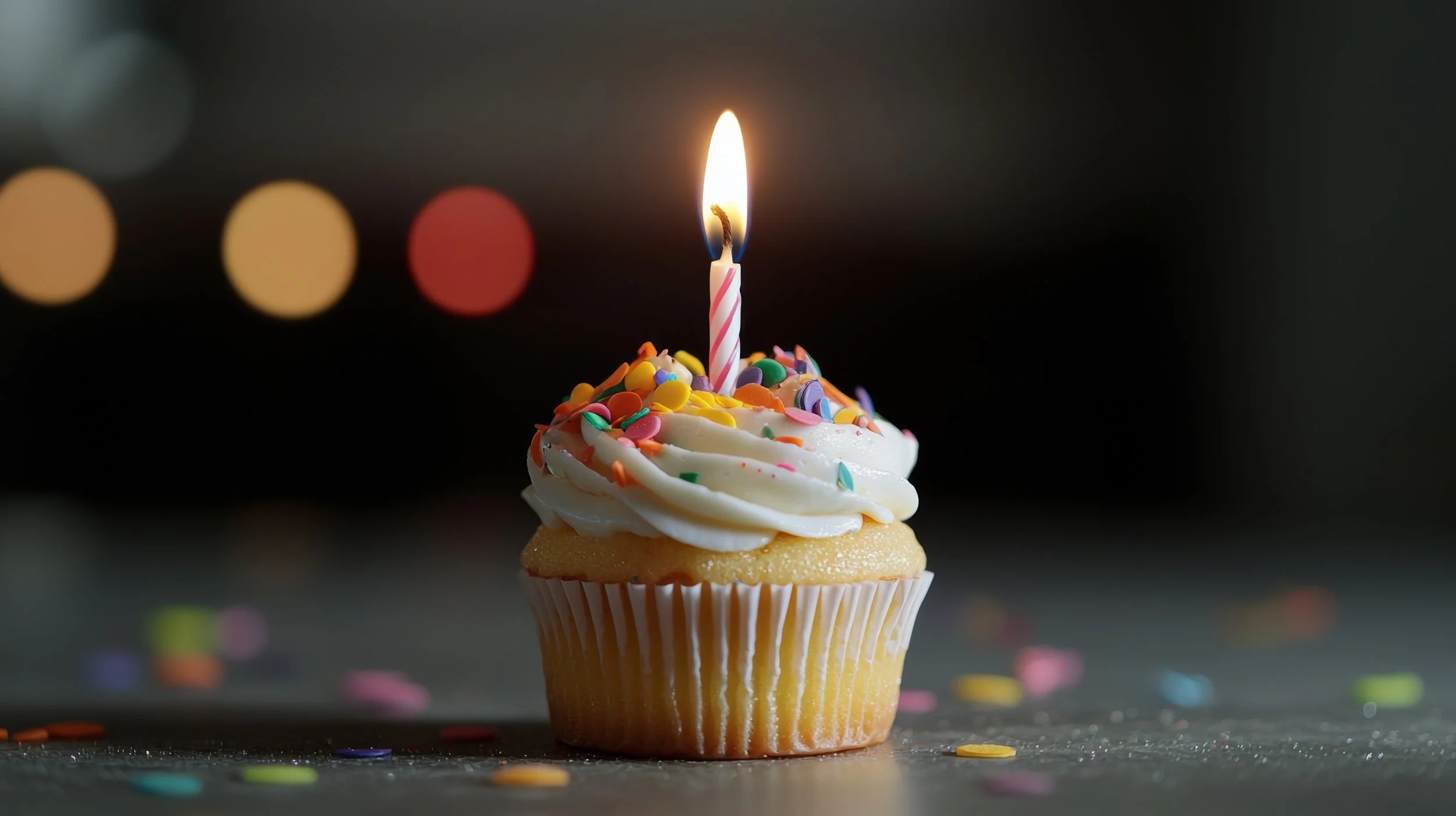 A birthday cake with white frosting, colorful sprinkles on top, and striped candles that are lit.