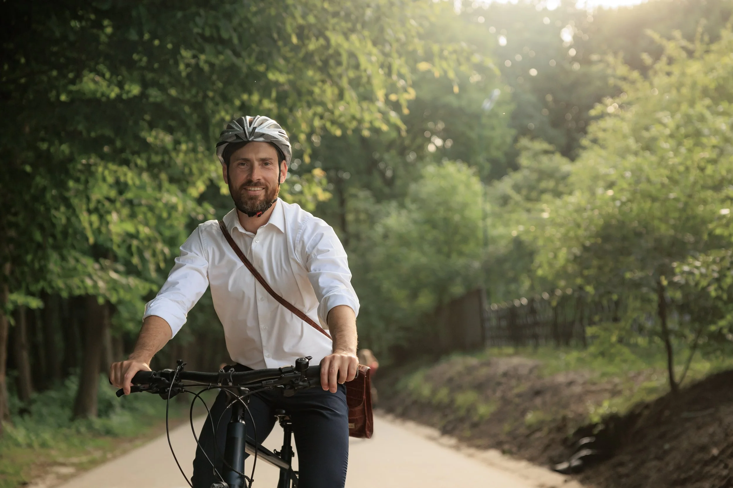 A man riding a bicycle on a tree-lined path during daytime, wearing a white shirt and a bike helmet, smiling at the camera.