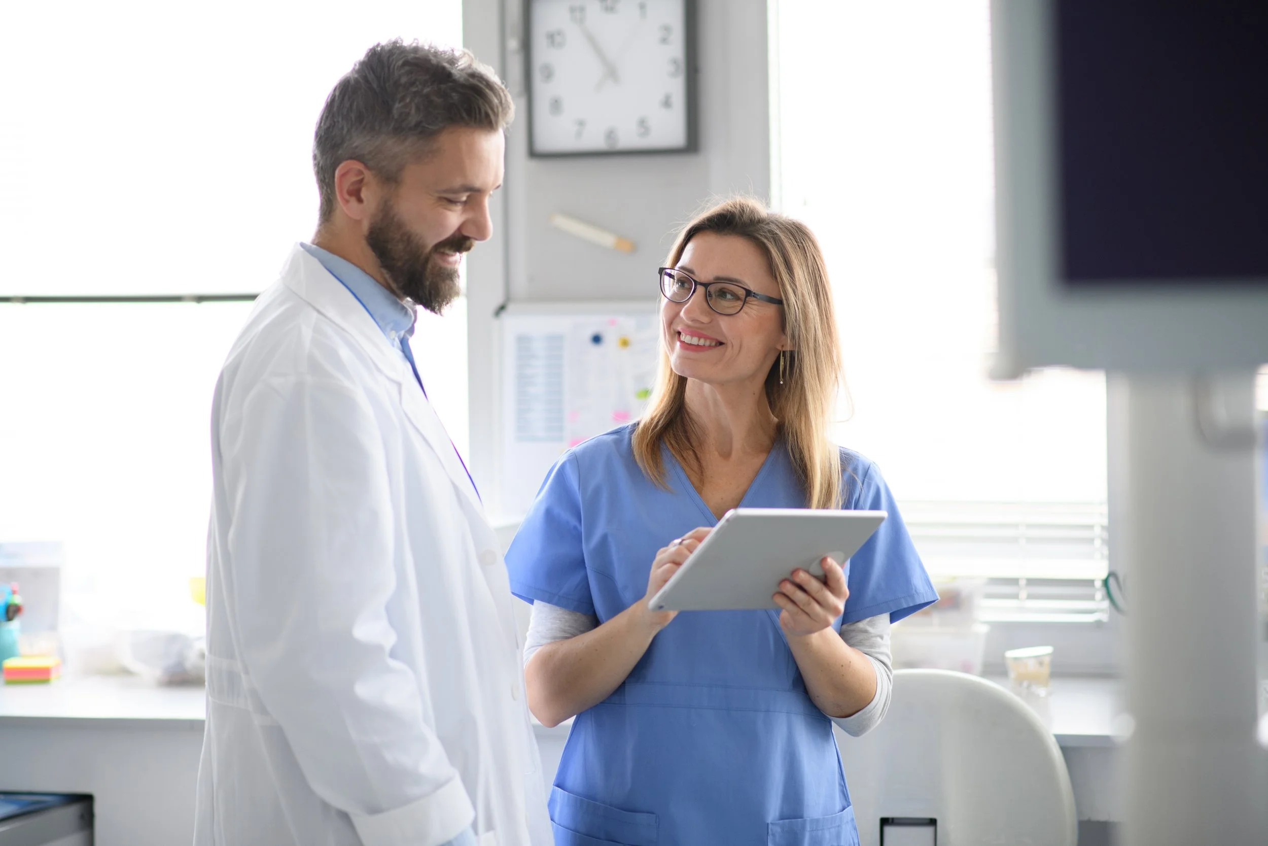 Two medical professionals, a man and a woman, are smiling and talking in a bright hospital or clinic setting. The woman is holding a tablet and wearing blue scrubs, while the man is in a white lab coat.