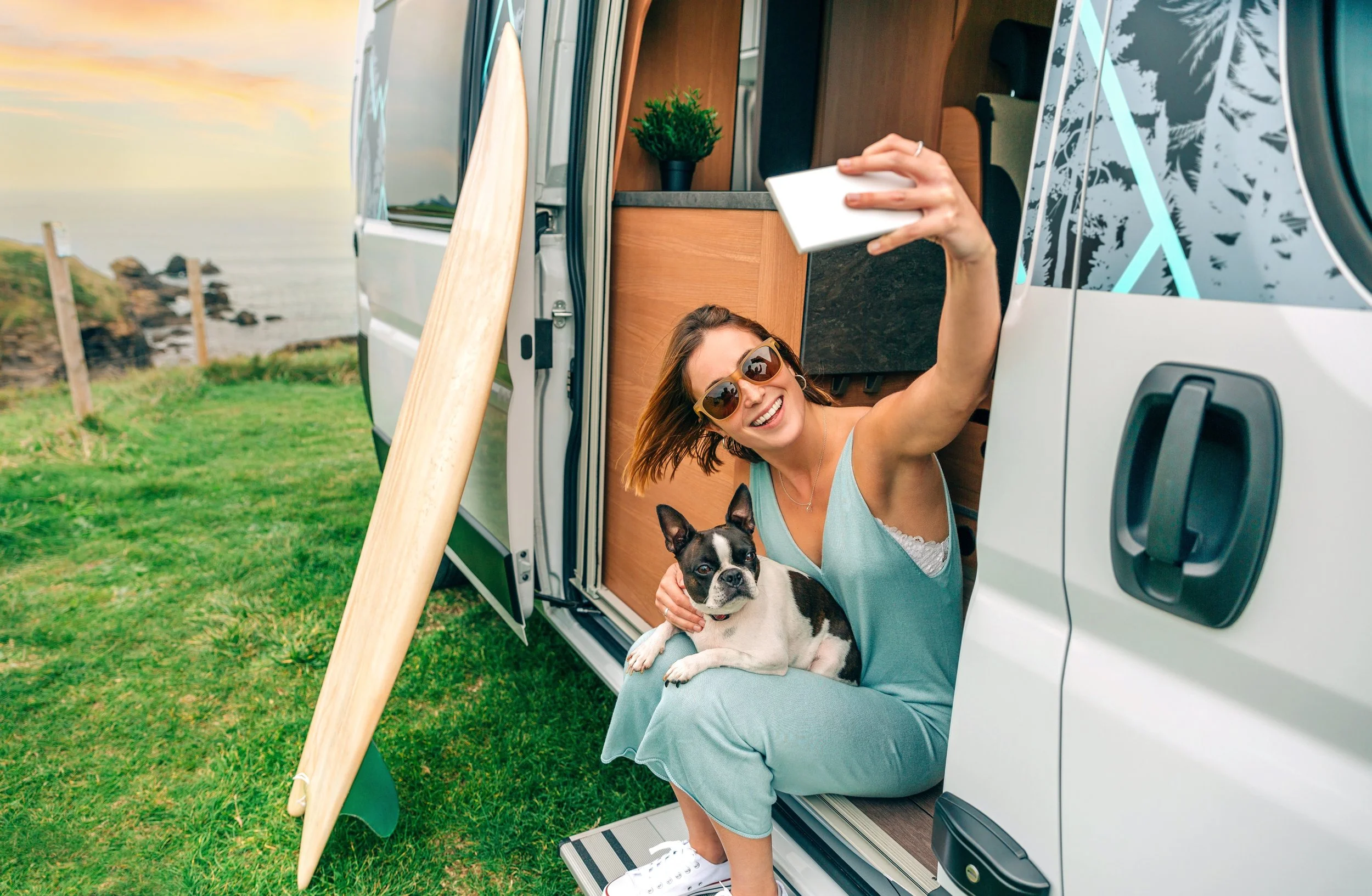 A woman with brown hair, wearing sunglasses, a turquoise outfit, and white sneakers, is sitting in the open doorway of a camper van. She is smiling and taking a selfie with a small black and white dog sitting on her lap. The camper van is parked outdoors on a grassy area near the coast, with a surfboard leaning against the van and a scenic view of the ocean and rocky shoreline in the background.