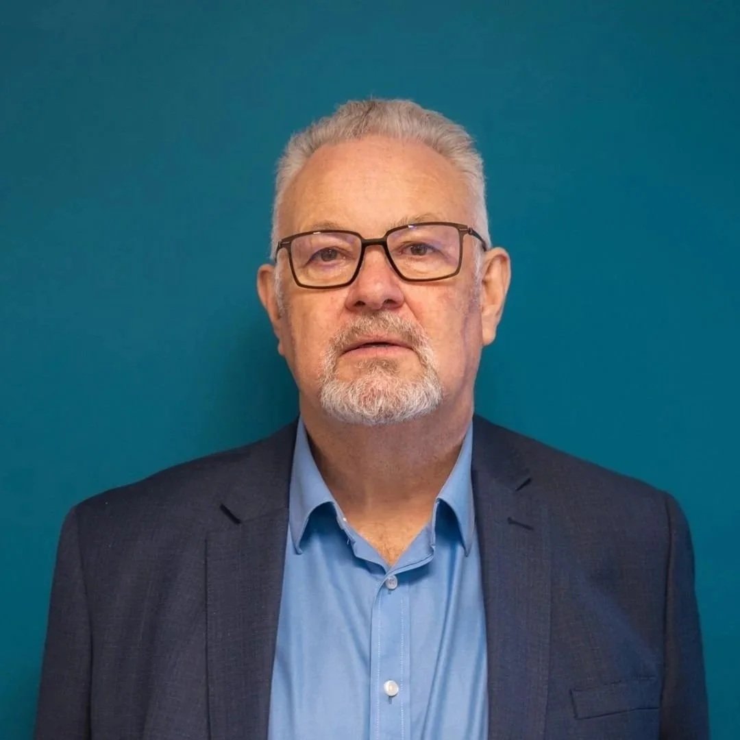 A middle-aged man with gray hair, beard, and glasses wearing a blue shirt and dark blazer against a teal background.