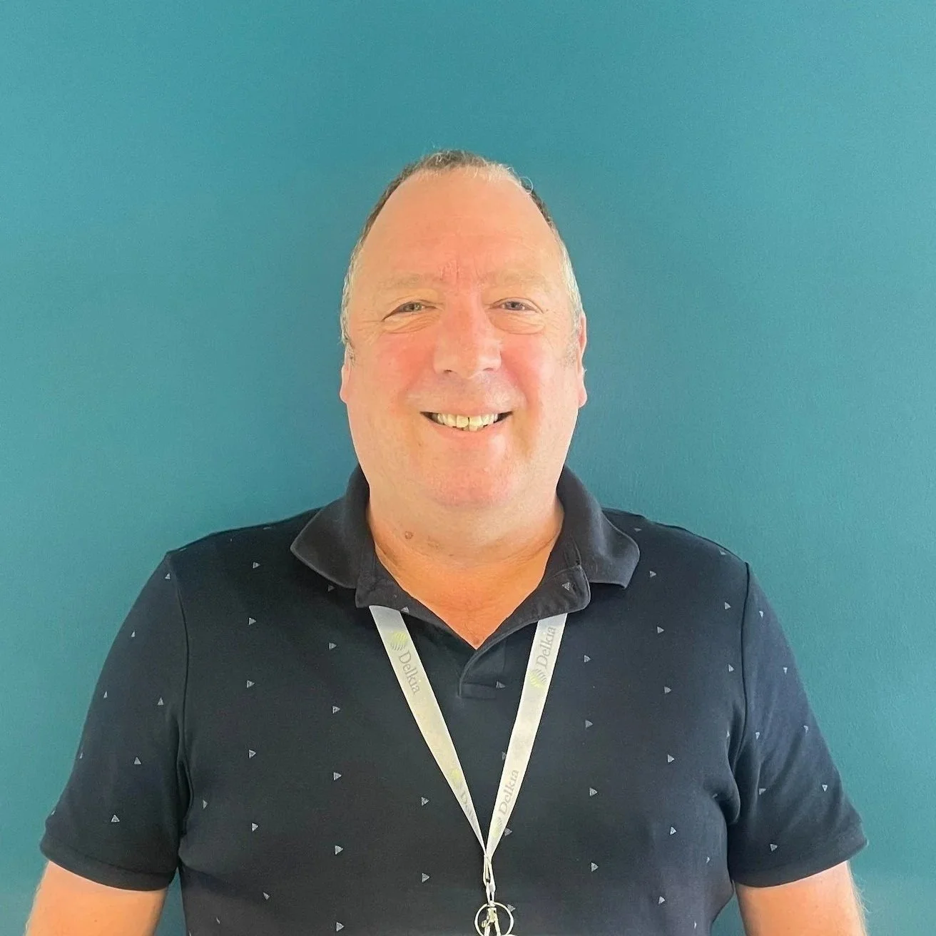 A smiling man with short hair wearing a black collared shirt with small white triangle patterns, standing against a teal wall, with a lanyard around his neck.