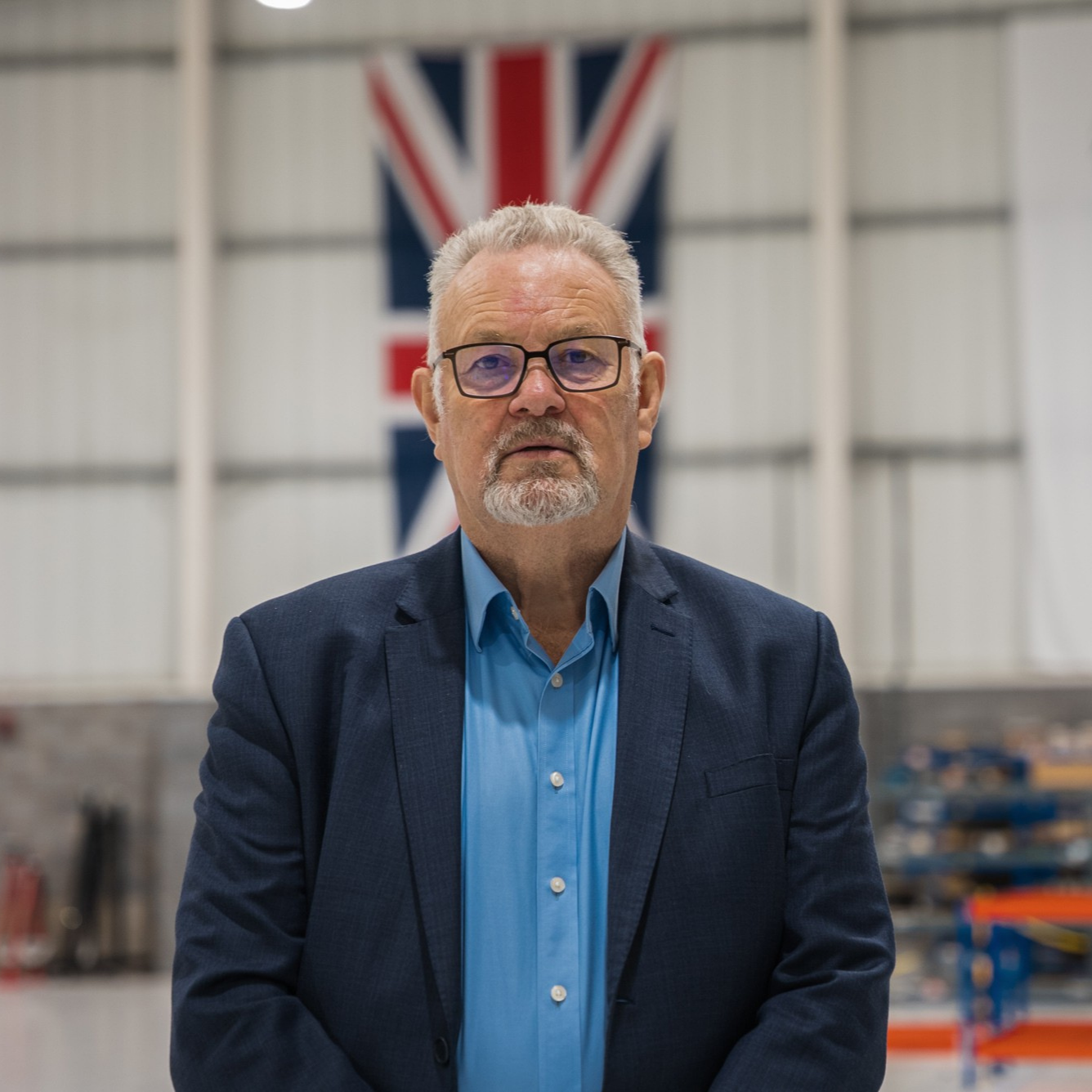 Man in blue shirt and blue suit, with glasses, white hair and a beard looking into the camera lens, a union flag hanging behind him.