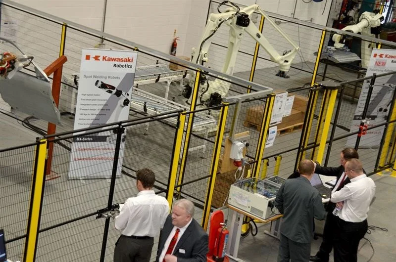 Robotic arms and machinery in an industrial setting, with four men in business attire discussing near a control panel.
