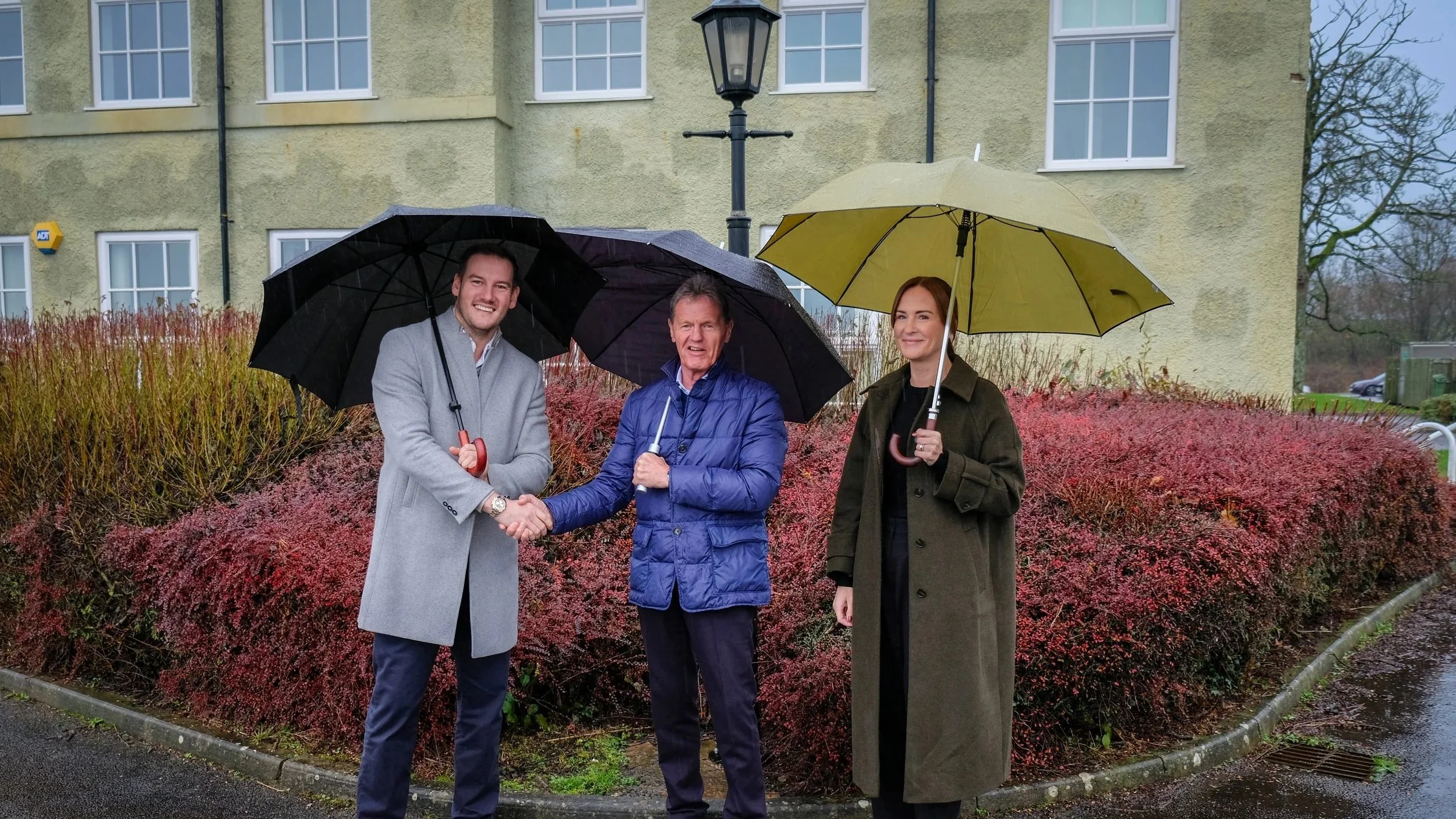 3 people in the rain with umbrellas, 2 men (shaking hands) and a lady - all smiling