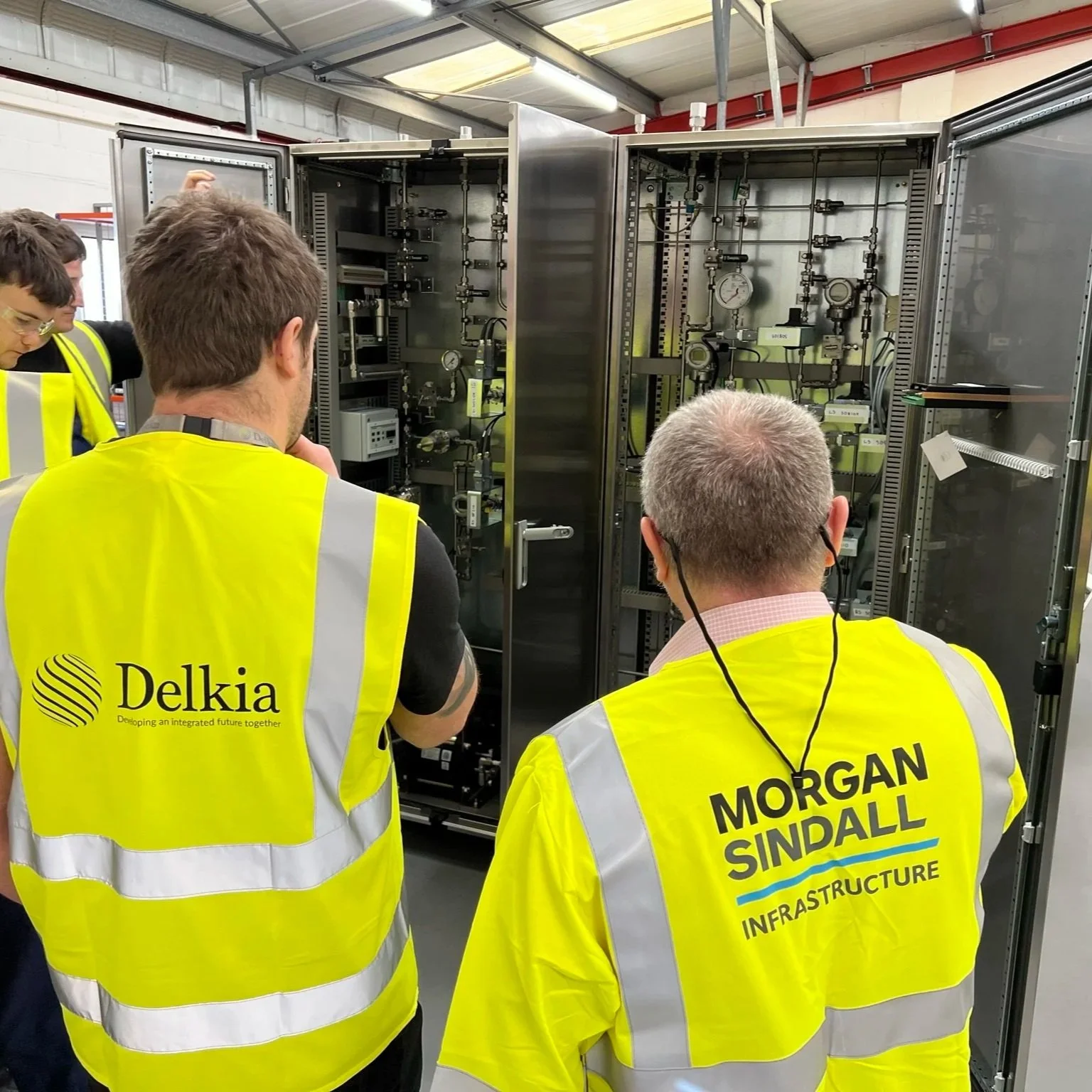 Group of people wearing yellow high-visibility vests inspecting large electrical or infrastructure equipment inside a facility.
