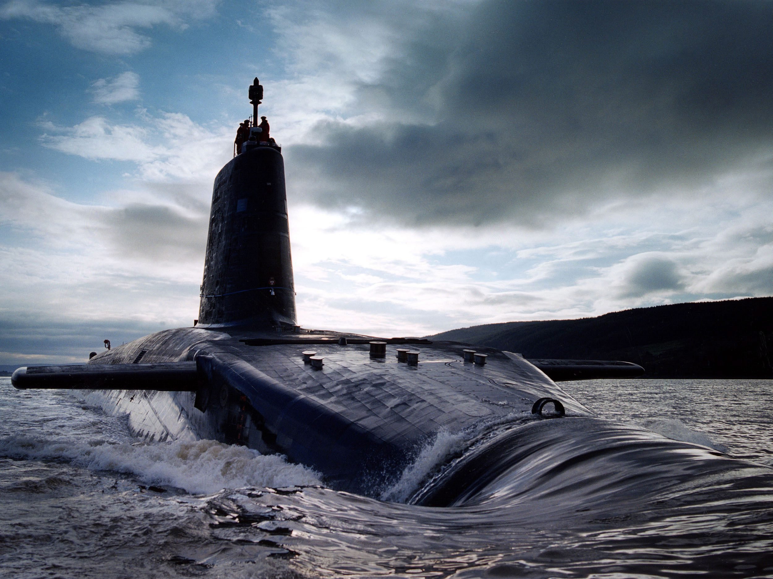 Grey British Navy sub against a cloudy blue sky as it sinks (or is rising) above/below sea level. 