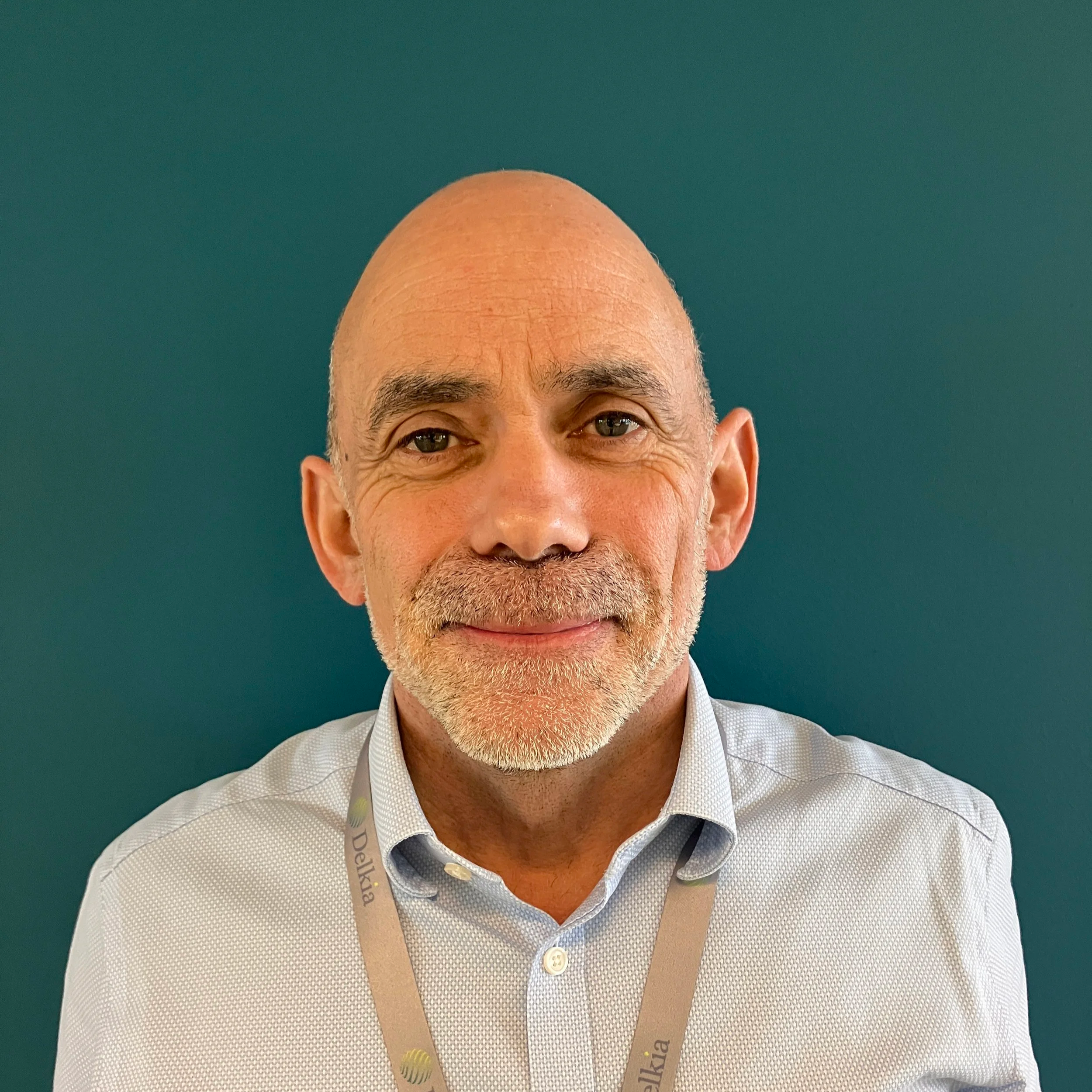 Portrait of a middle-aged man with a bald head, light beard, and mustache, wearing a light-colored collared shirt and a lanyard against a teal background.